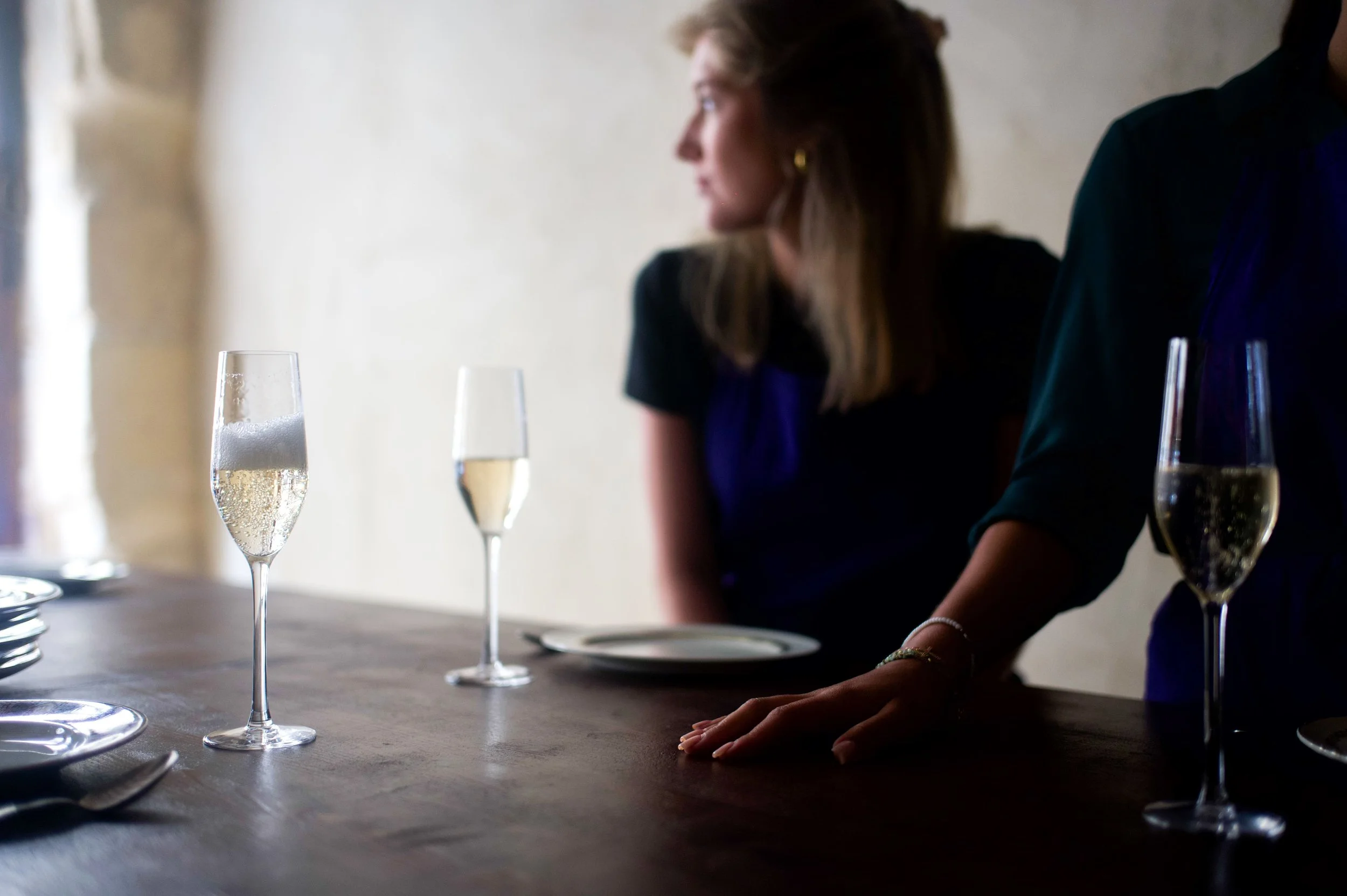 Verres de champagne sur une table en bois avec deux femmes à l'arrière, une de profil et une autre en partie visible, dans un décor intérieur