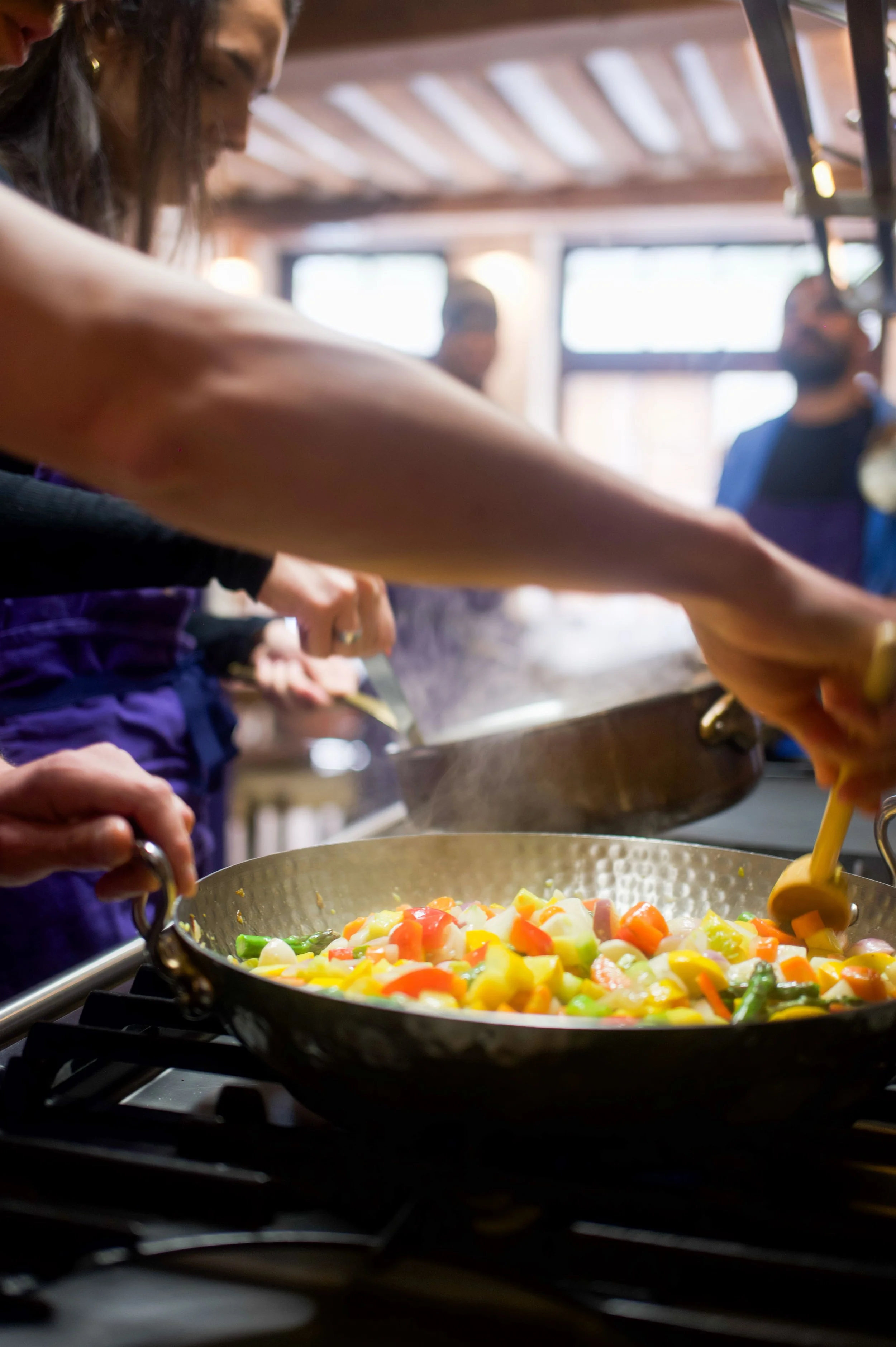 Groupe de personnes cuisinant ensemble dans une cuisine, en train de préparer un plat de légumes sautés dans une poêle en cuivre