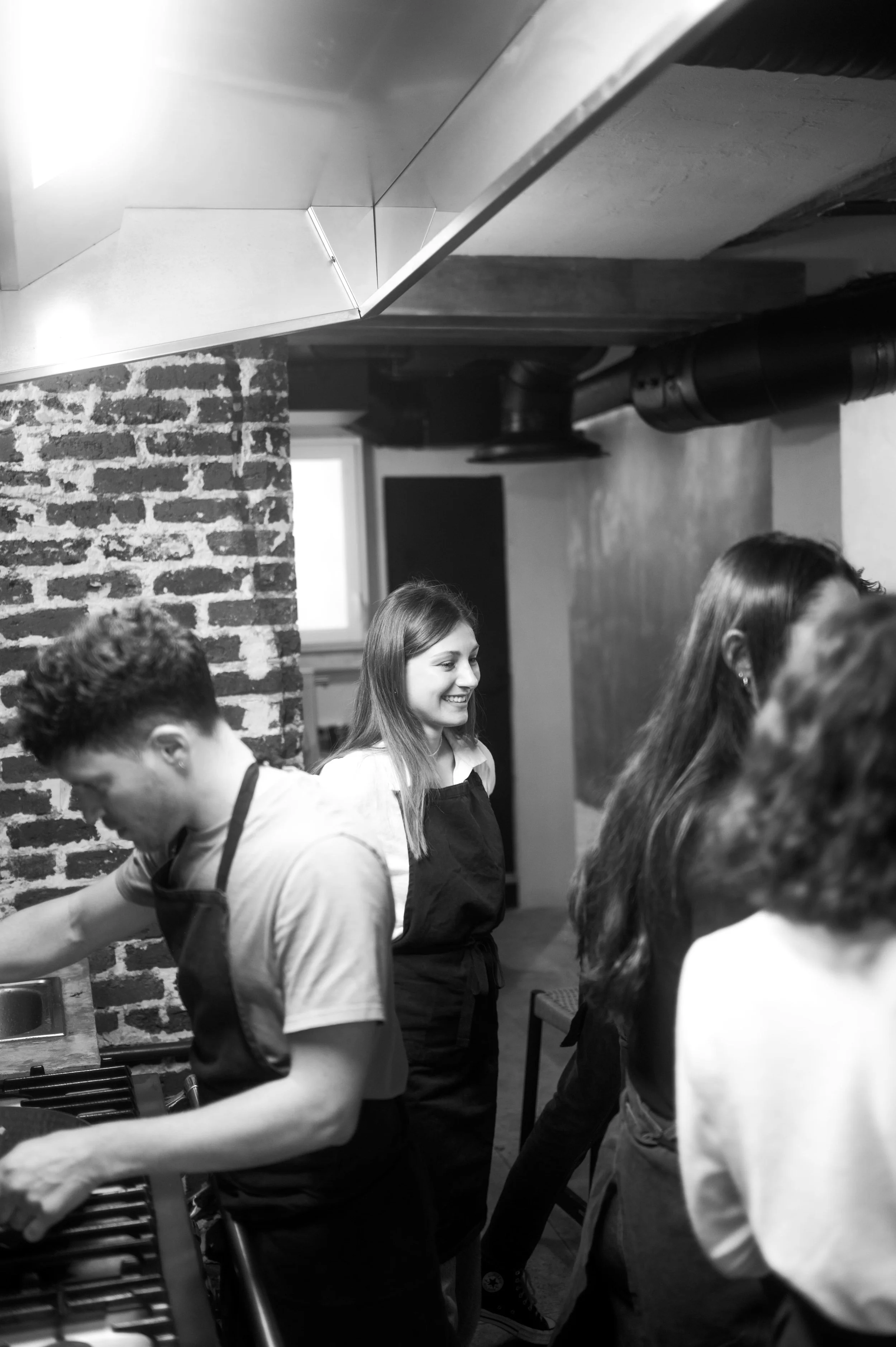 Groupe de jeunes personnes en cuisine, portant des tabliers, en train de cuisiner et de discuter, dans une ambiance chaleureuse et conviviale.