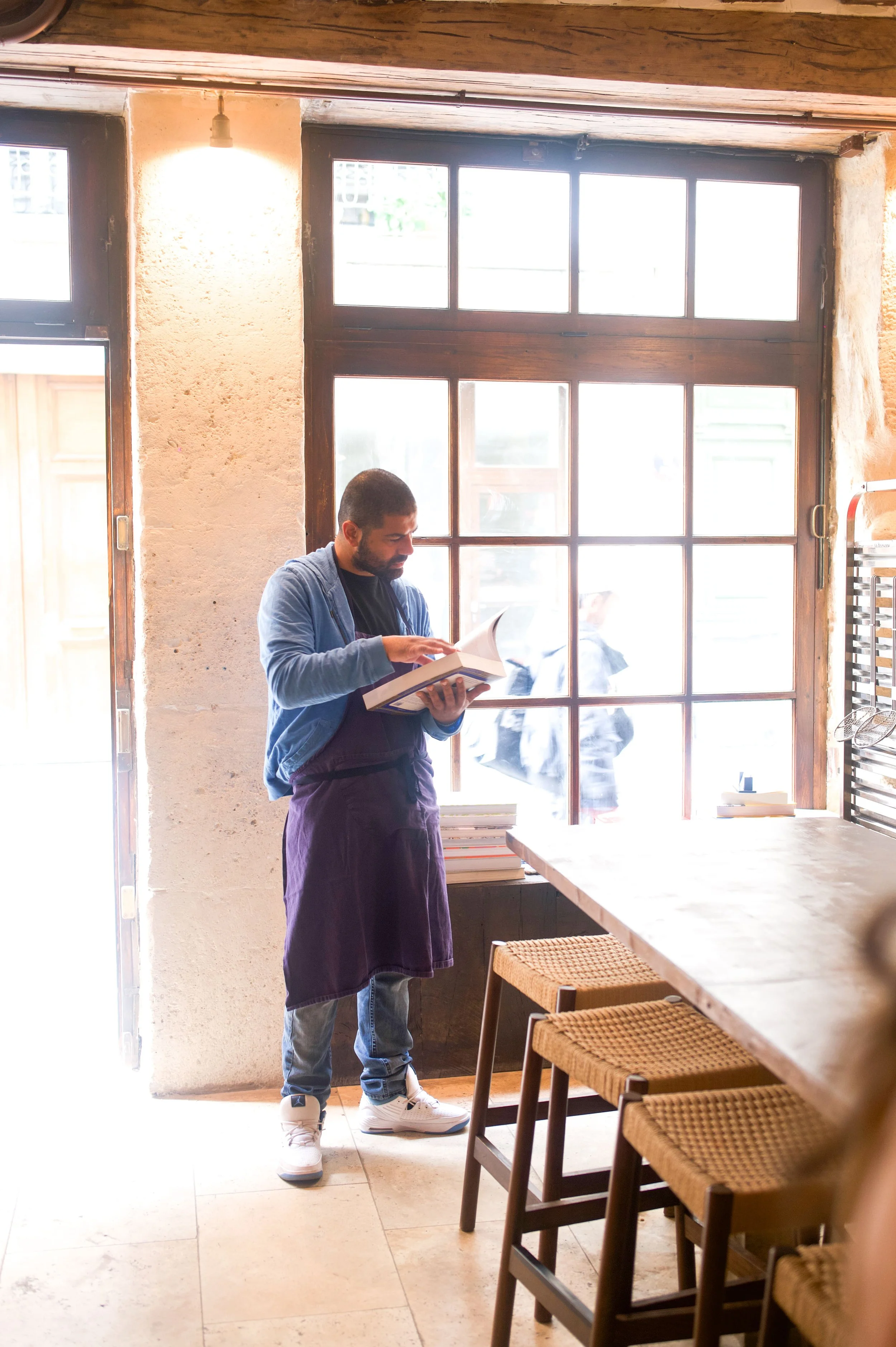 Un homme debout à côté d'une table en bois, tenant un livre ouvert et lisant, dans un intérieur lumineux avec une grande fenêtre en bois, style rustique.