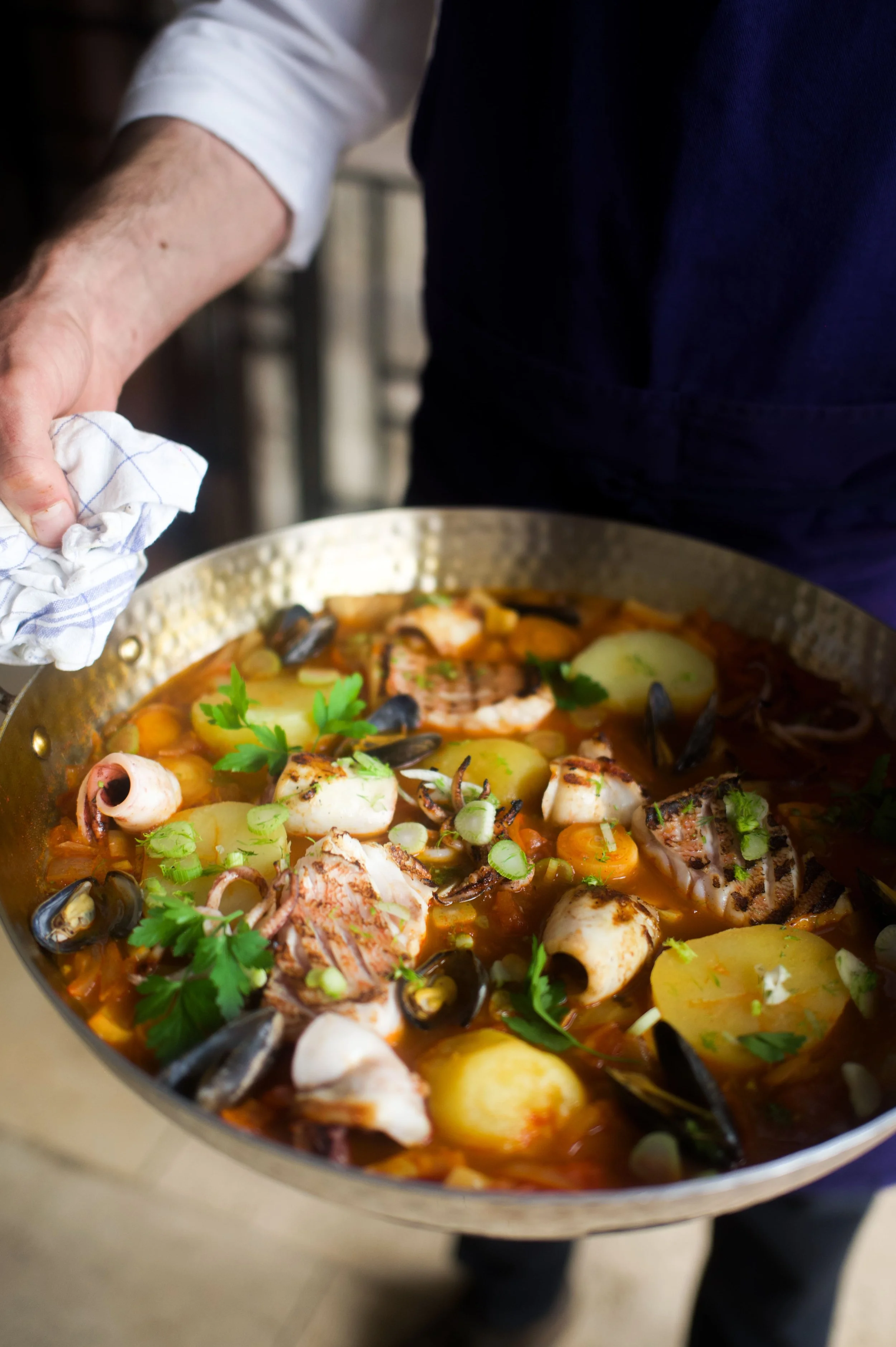 Une personne tient une poêle contenant un plat de fruits de mer, comprenant des morceaux de poisson, des moules, des pommes de terre, de la sauce tomate et des herbes fraîches.
