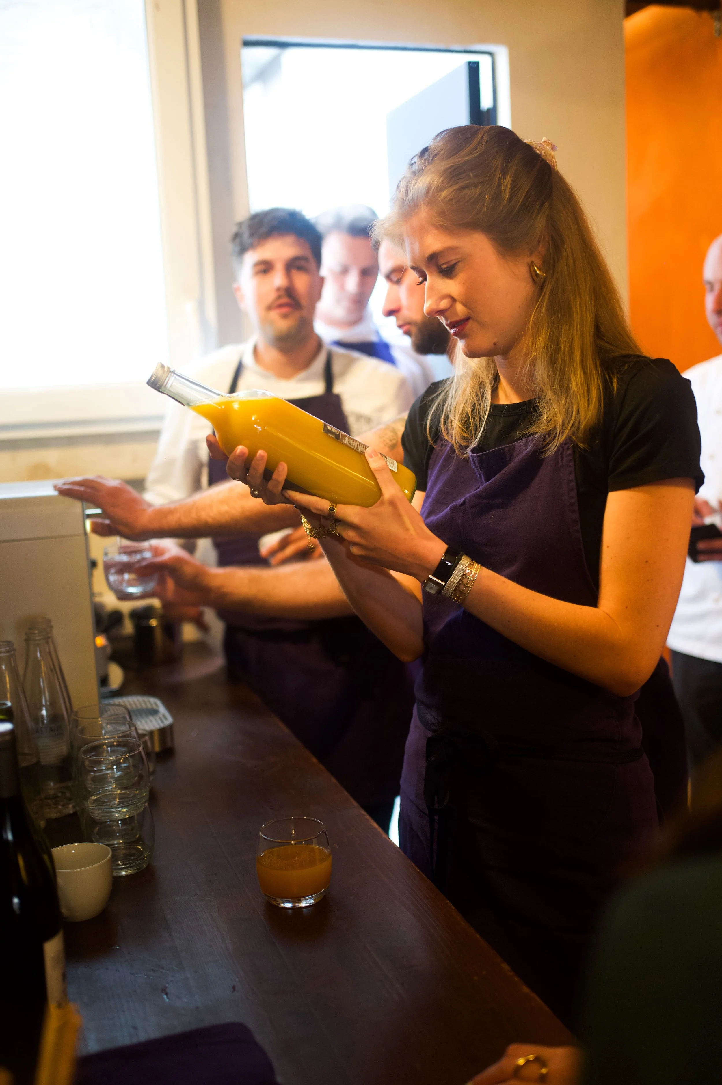 Une femme regarde une bouteille de jus d'orange pendant que d'autres personnes derrière elle attendent ou préparent du café, en intérieur