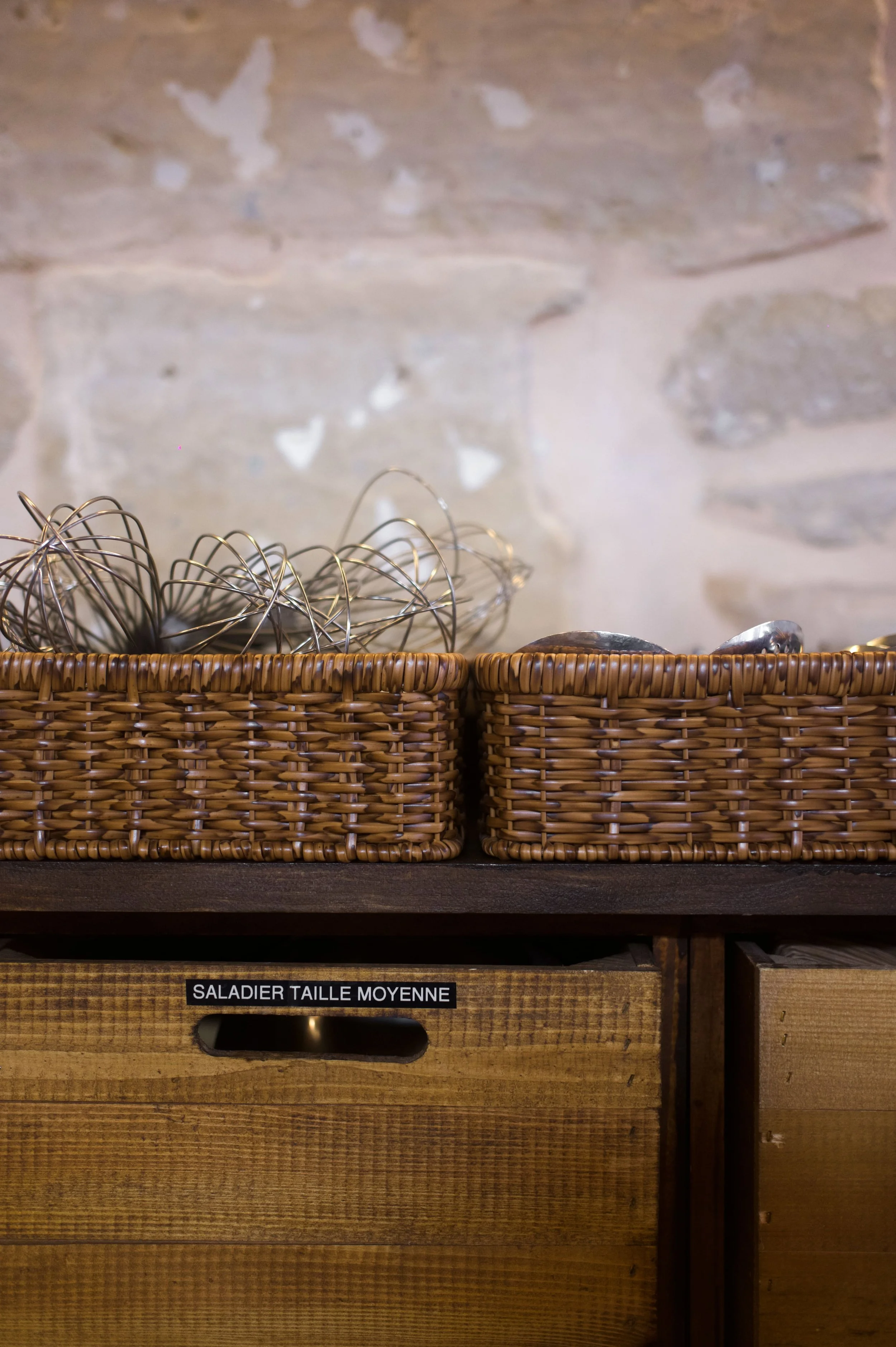 Rattan diapers with metal whisks and spoons in baskets, on a wooden cabinet labeled "Salad Bowl Medium Size". Stone wall in background.