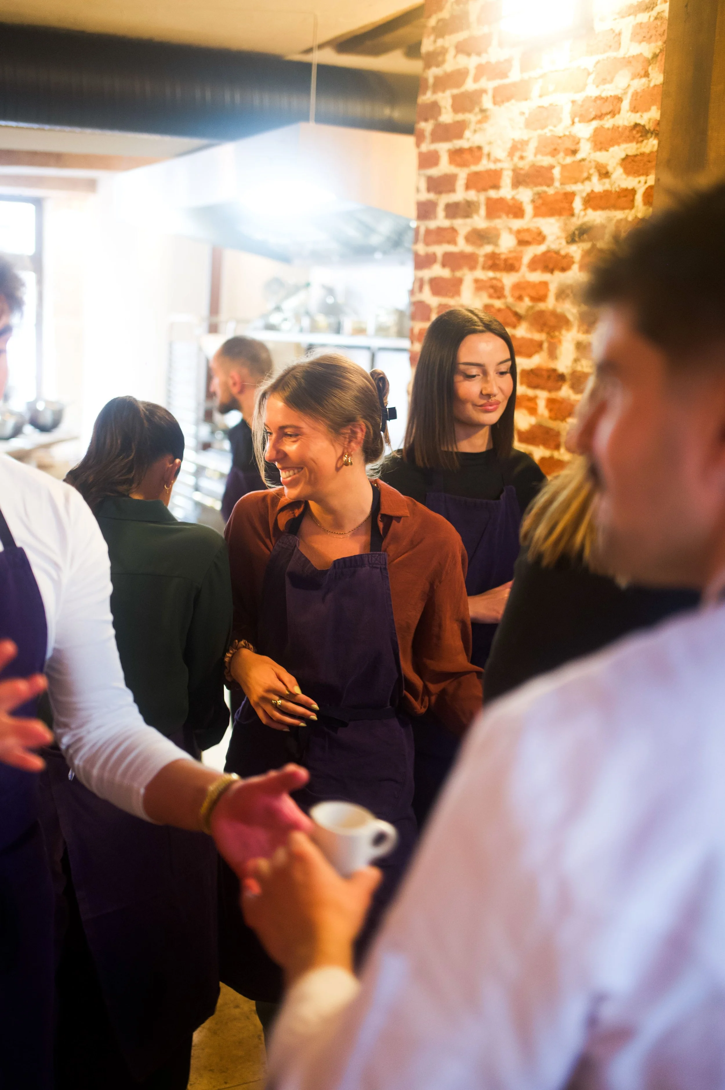 Groupe de personnes travaillant en cuisine ou dans un café, discutant et souriant, avec un mur en briques en arrière-plan.