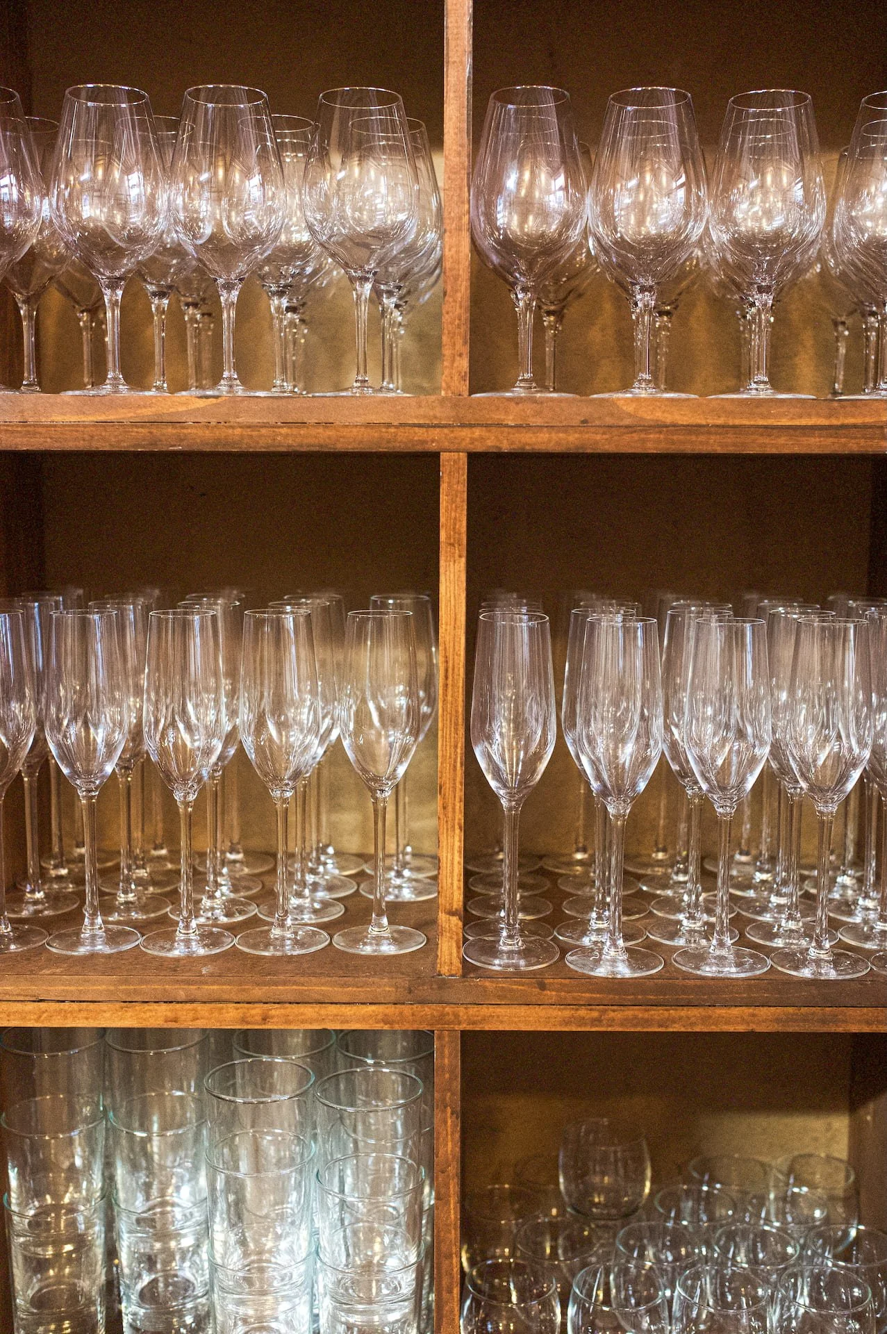 Glass glasses of various shapes and sizes arranged on a wooden shelf.
