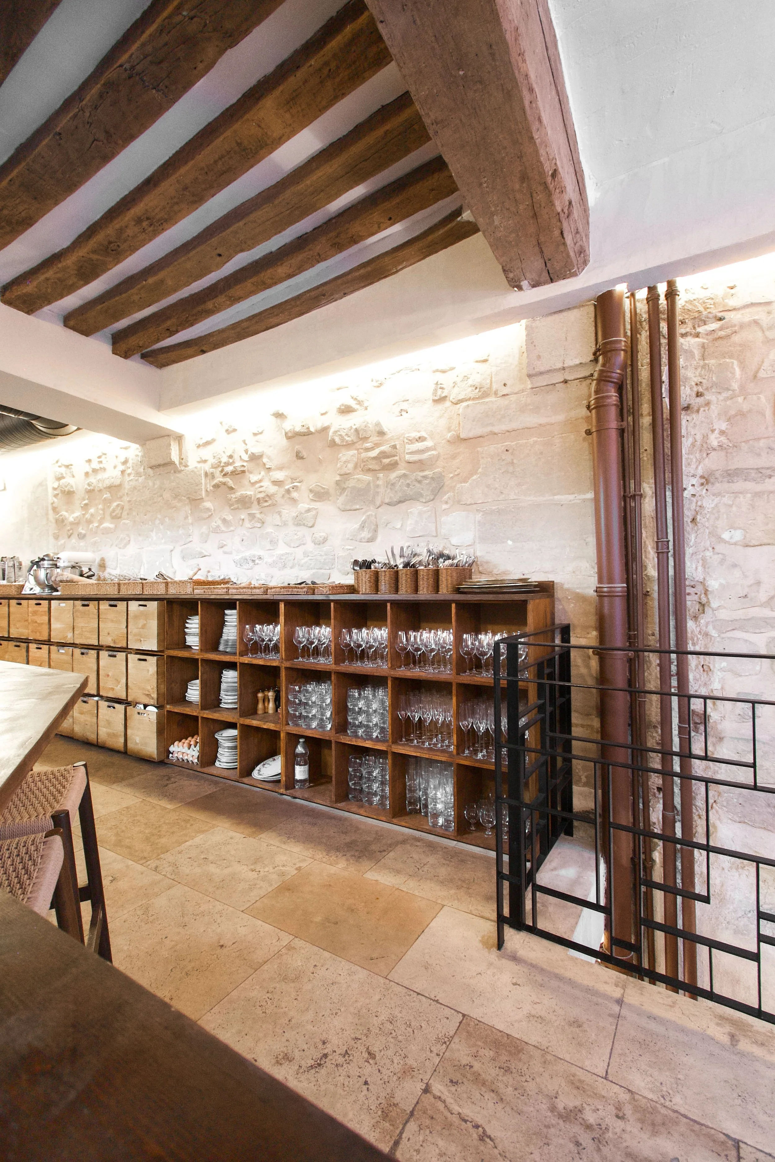 Wooden shelf with wine glasses, plates and utensils in a rustic kitchen with stone walls and wood-beamed ceiling.