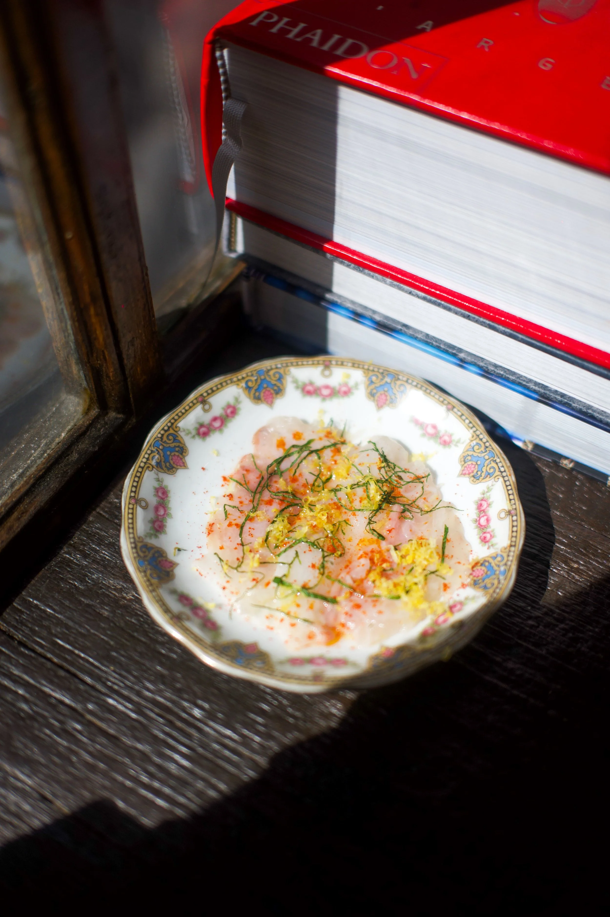 Une petite assiette décorative avec un plat de carpaccio de poisson garni de zestes de citron et d'herbes, placée sur une surface en bois à côté d'un livre.