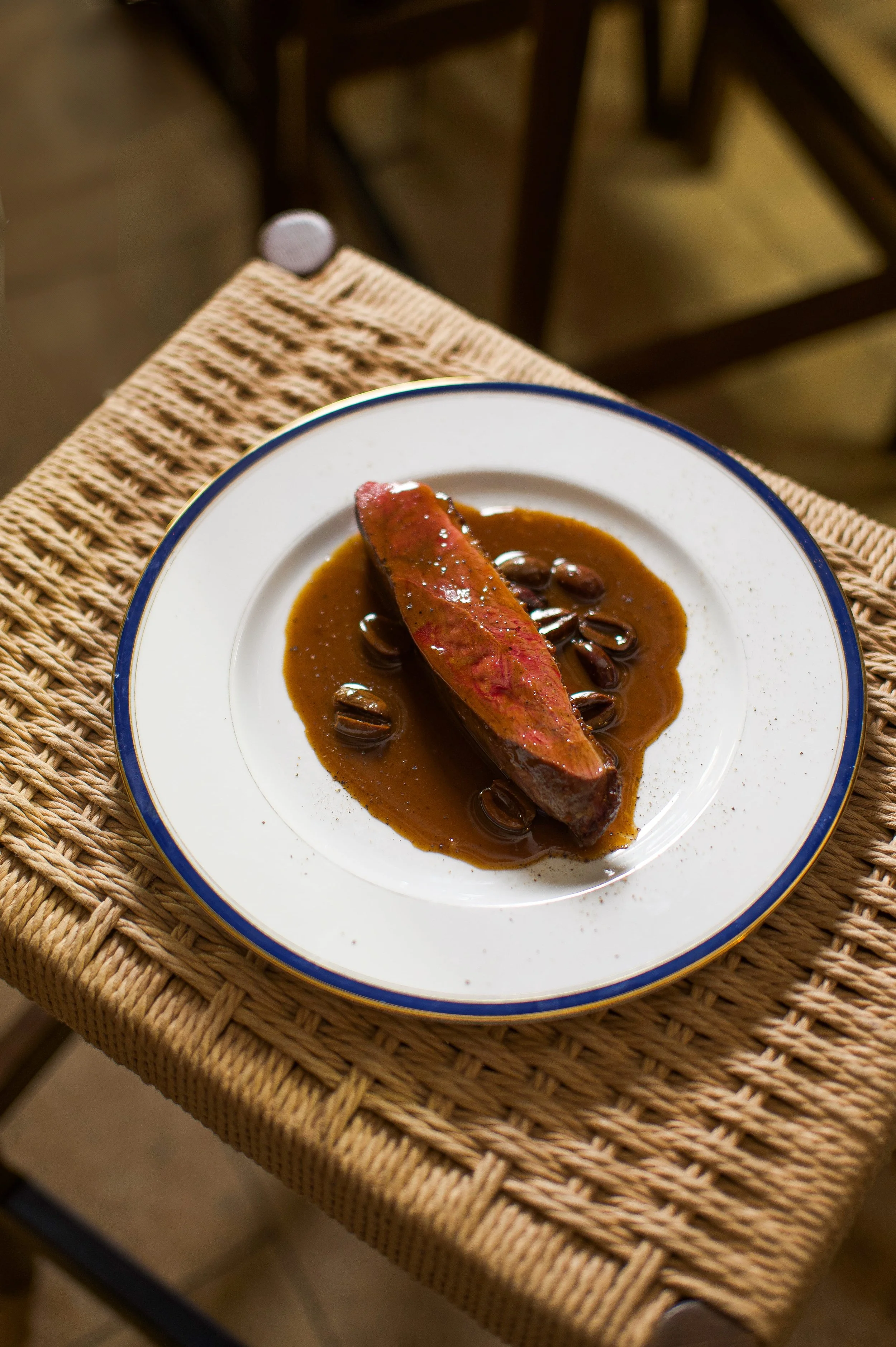 Plat de viande avec sauce et garniture sur une assiette blanche avec bord bleu, placé sur un tabouret en rotin.