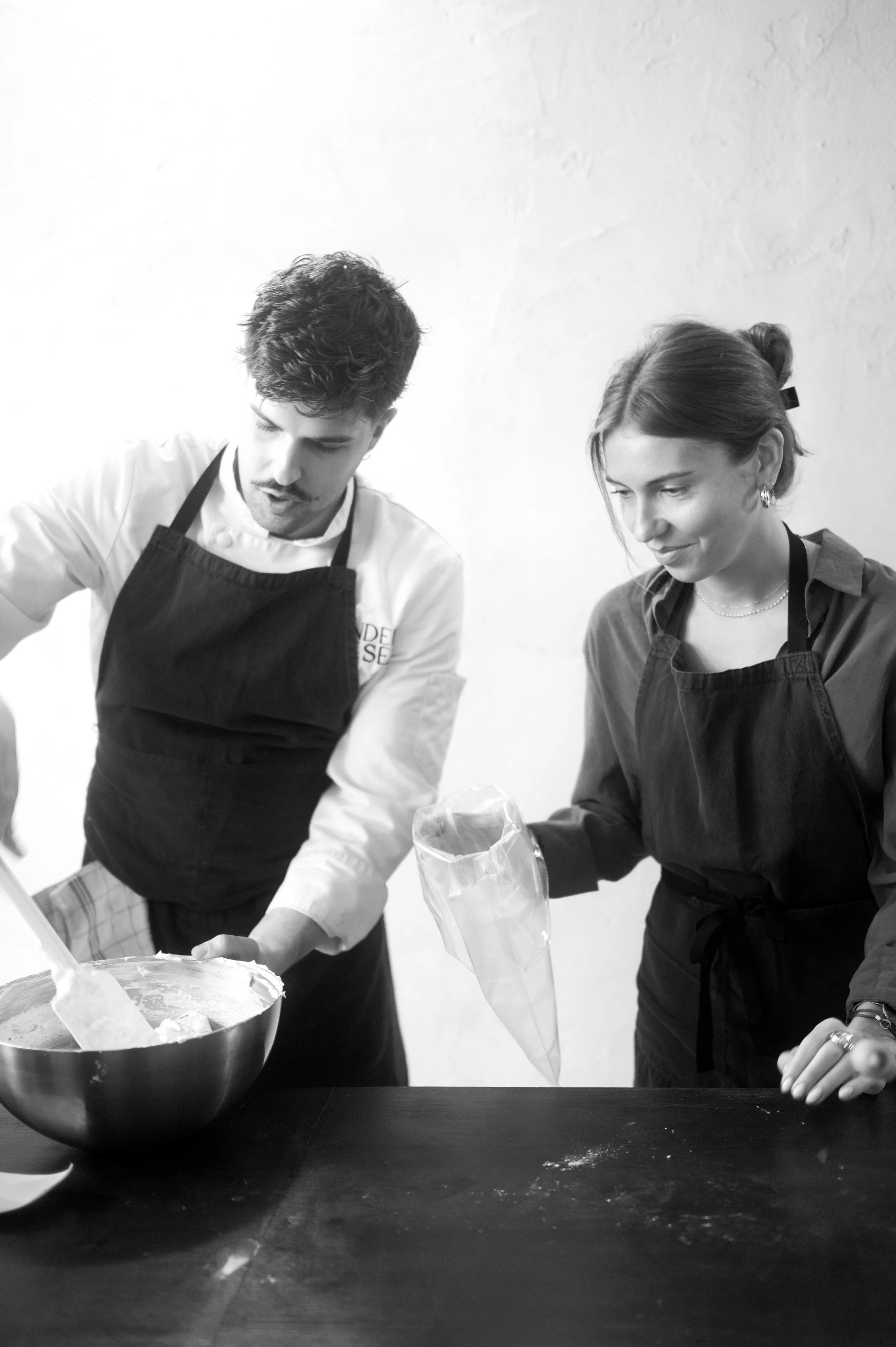 Deux personnes en tablier noir, un homme et une femme, préparant quelque chose en cuisine, avec un bol et un sac en plastique sur une table.