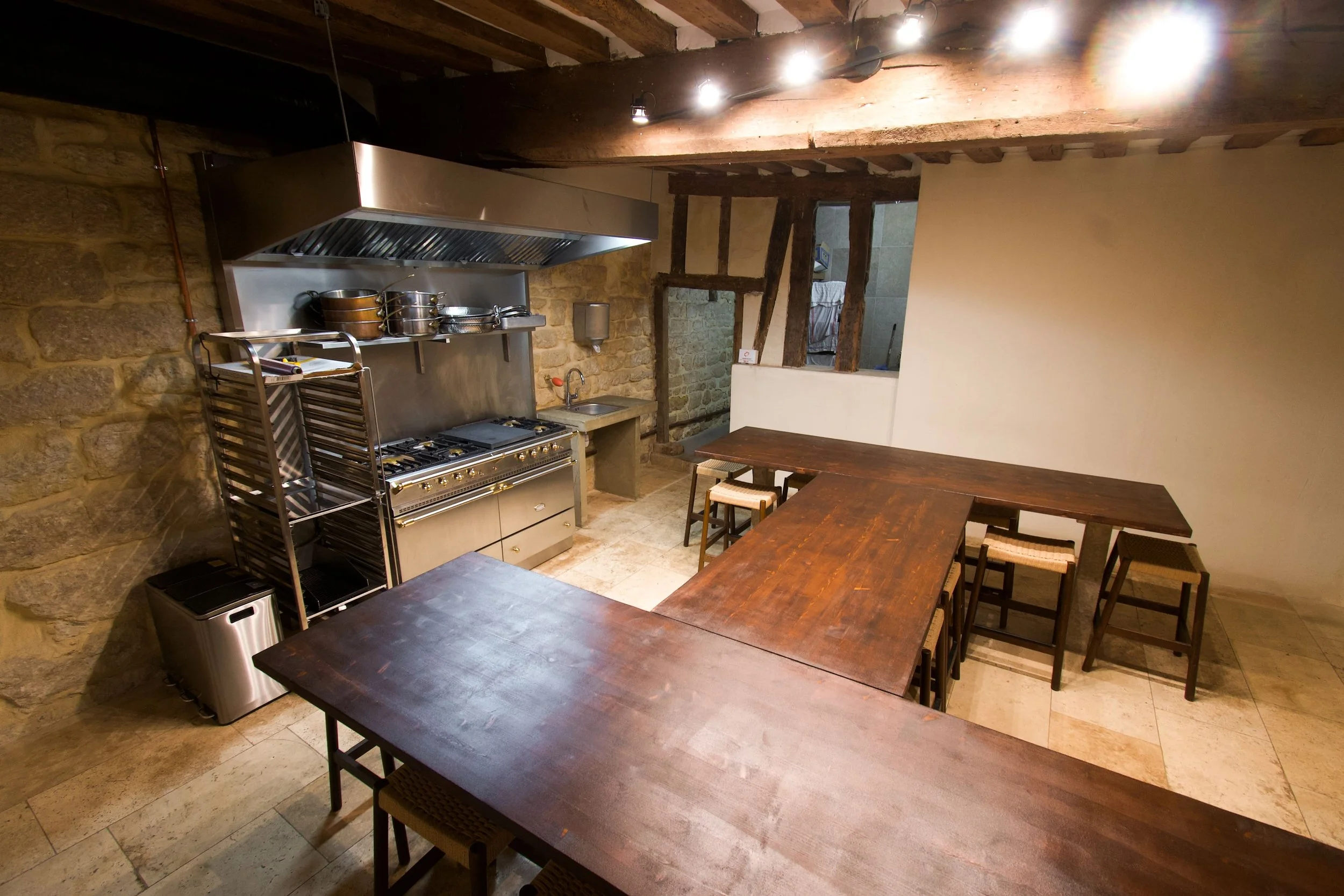 Restaurant kitchen with wooden tables, gas stove, stainless steel shelves, stone and wood walls, and pendant lighting.