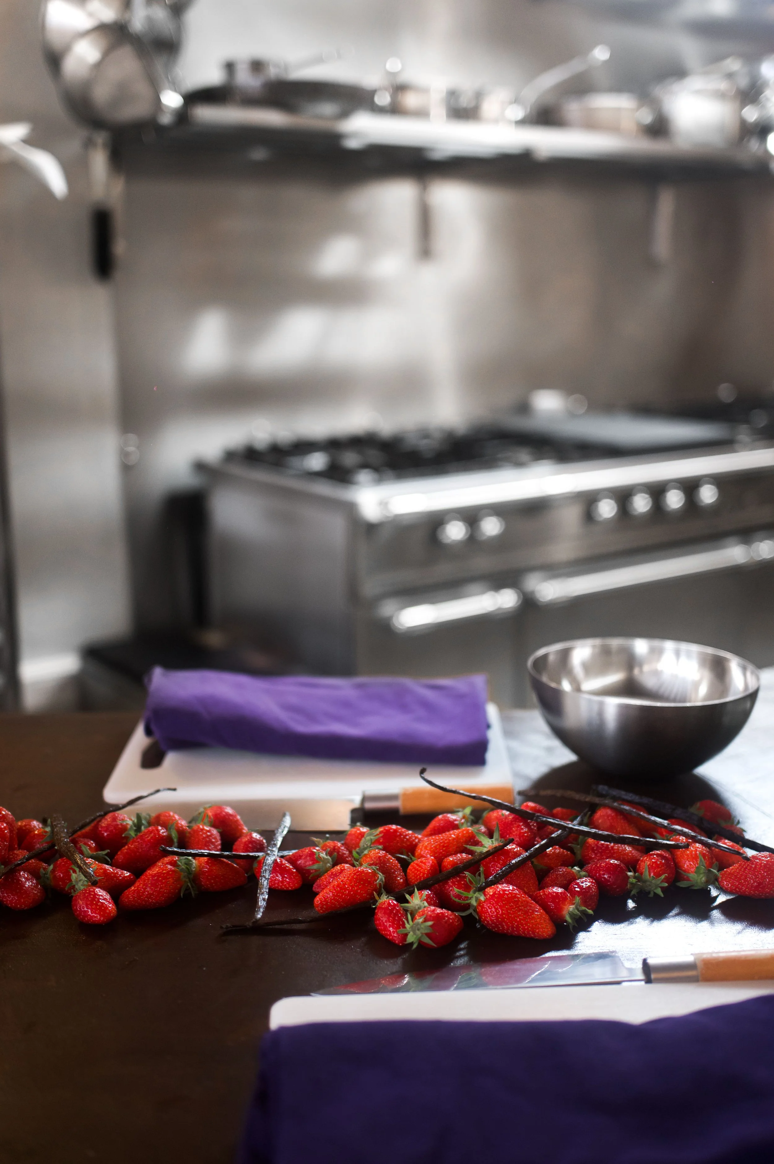 Fraises fraîches disposées sur une table de cuisine, avec un bol en acier inoxydable et des outils de cuisine, dans une cuisine professionnelle en arrière-plan.