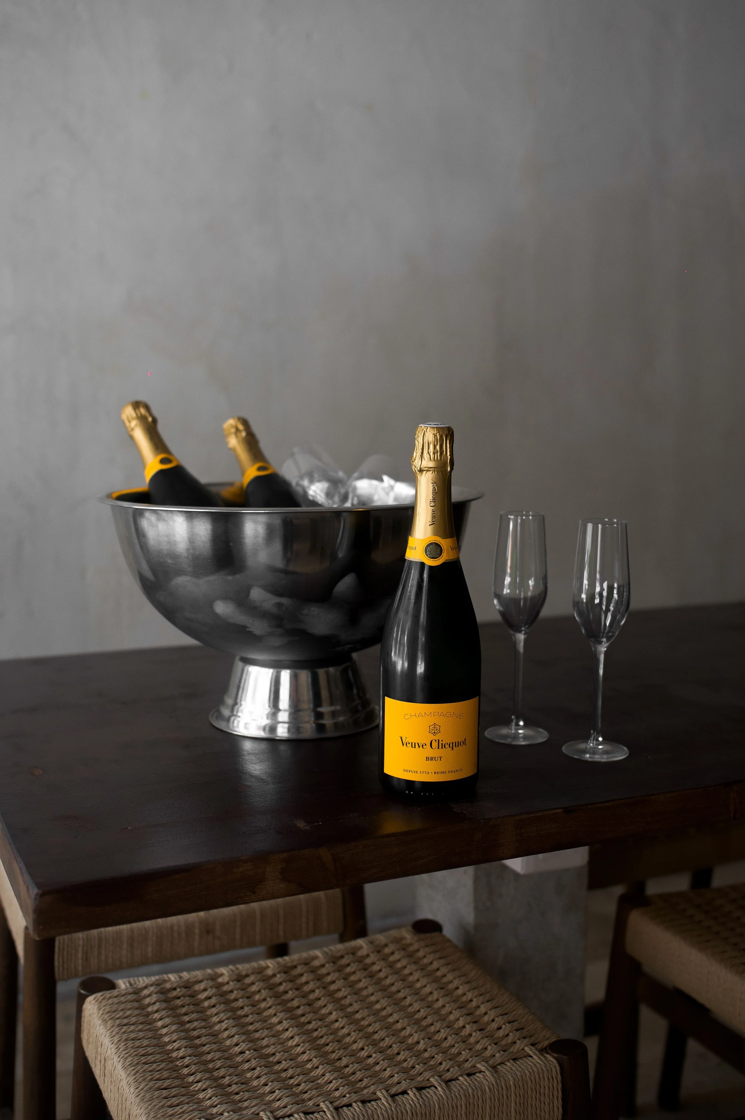 Une bouteille de champagne Veuve Clicquot devant deux flûtes à champagne vides sur une table en bois, avec une glacière contenant plusieurs autres bouteilles de champagne et des glaçons.