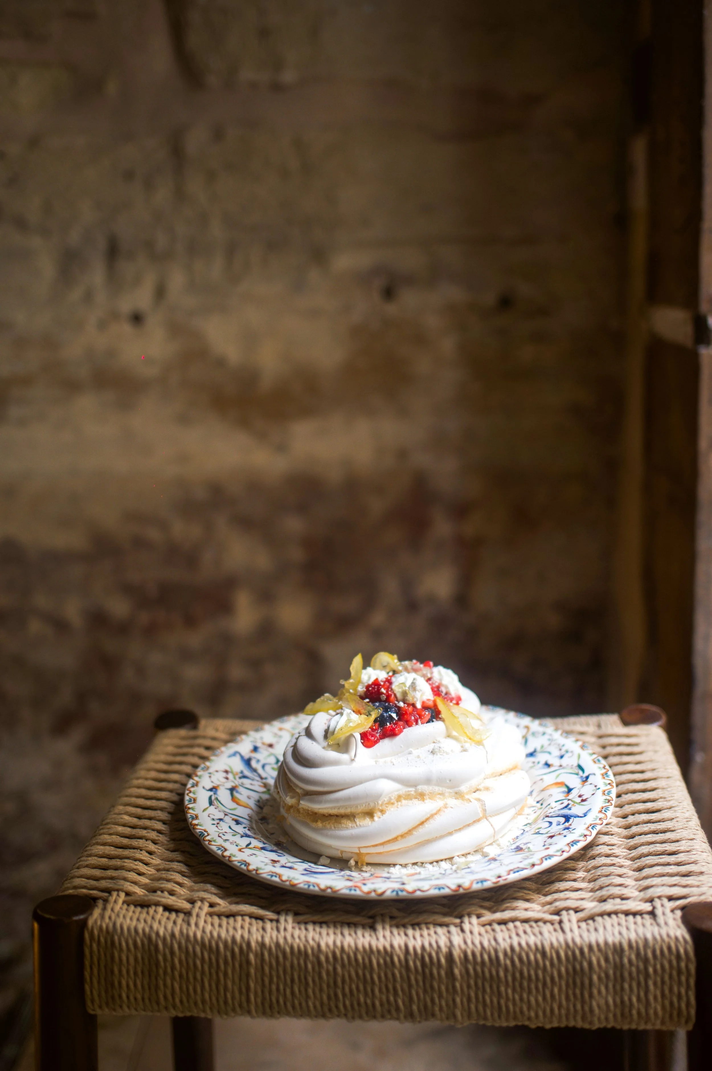 Une meringue garnie de chantilly, fruits rouges et zestes de citron, servie sur une assiette en céramique avec un motif coloré, placée sur un petit tabouret en osier.