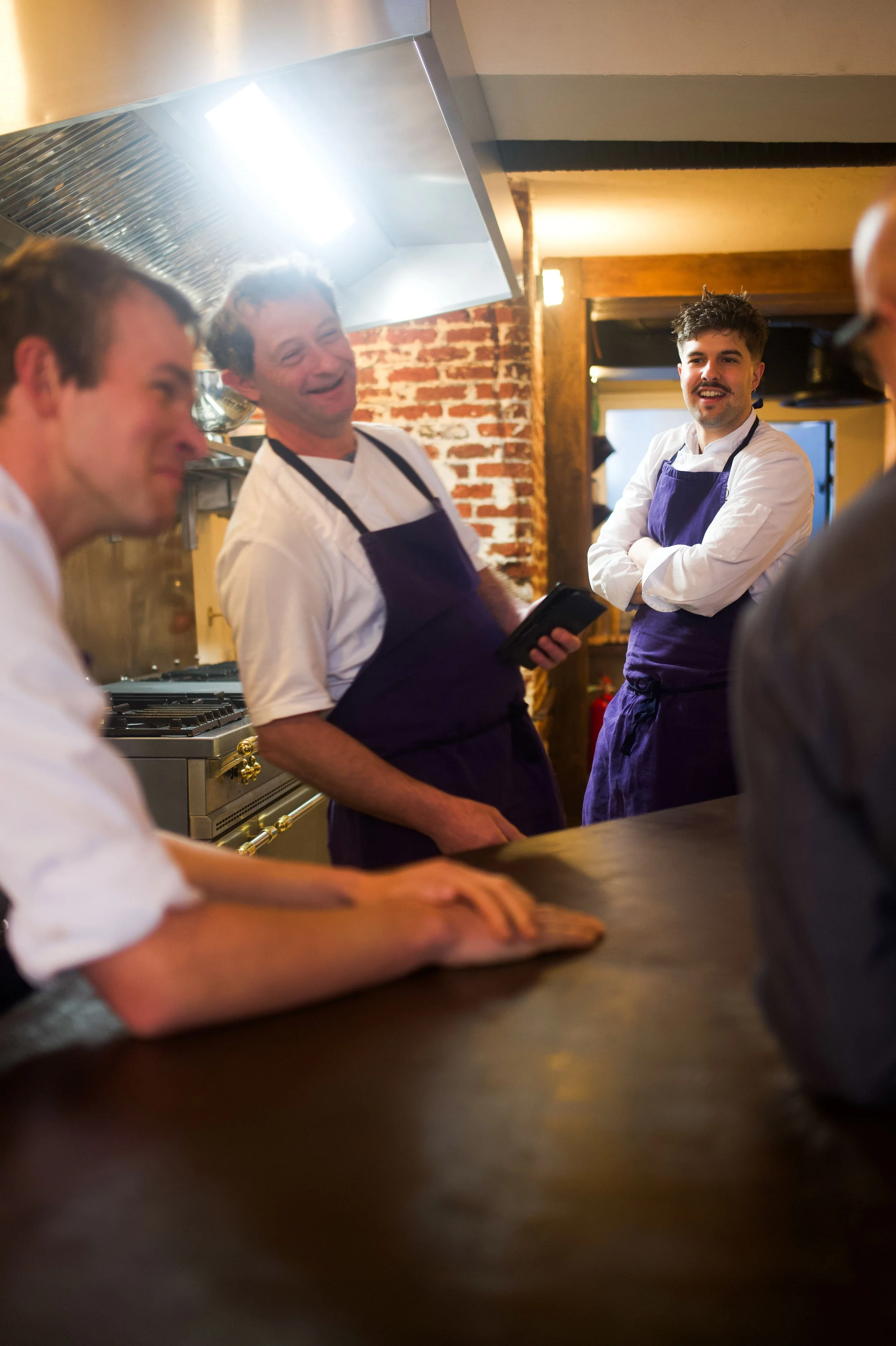 Quatre chefs cuisiniers en train de discuter dans une cuisine de restaurant, deux d'entre eux portent un tablier violet et un portent un appareil électronique dans la main, un autre a la main posée sur le comptoir en bois.