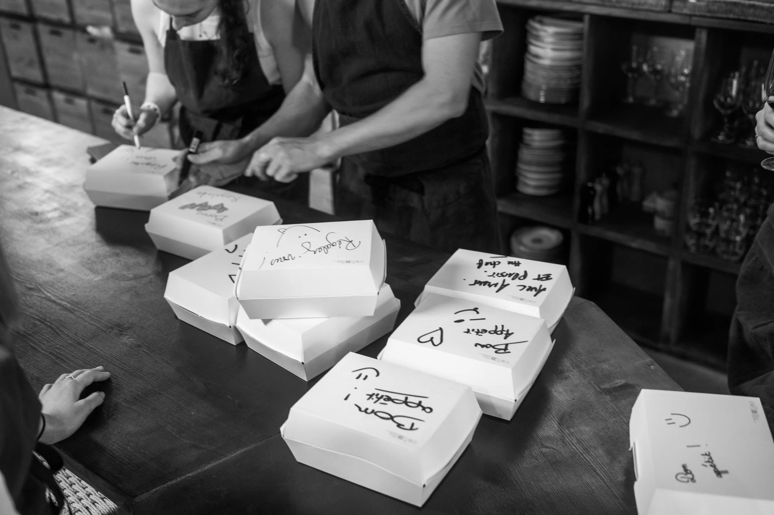 Gros plan sur une table en bois avec plusieurs boîtes à emporter empilées et signées à la main, avec des personnes en train de préparer ou de signer des emballages, dans une ambiance de cuisine ou de restaurant