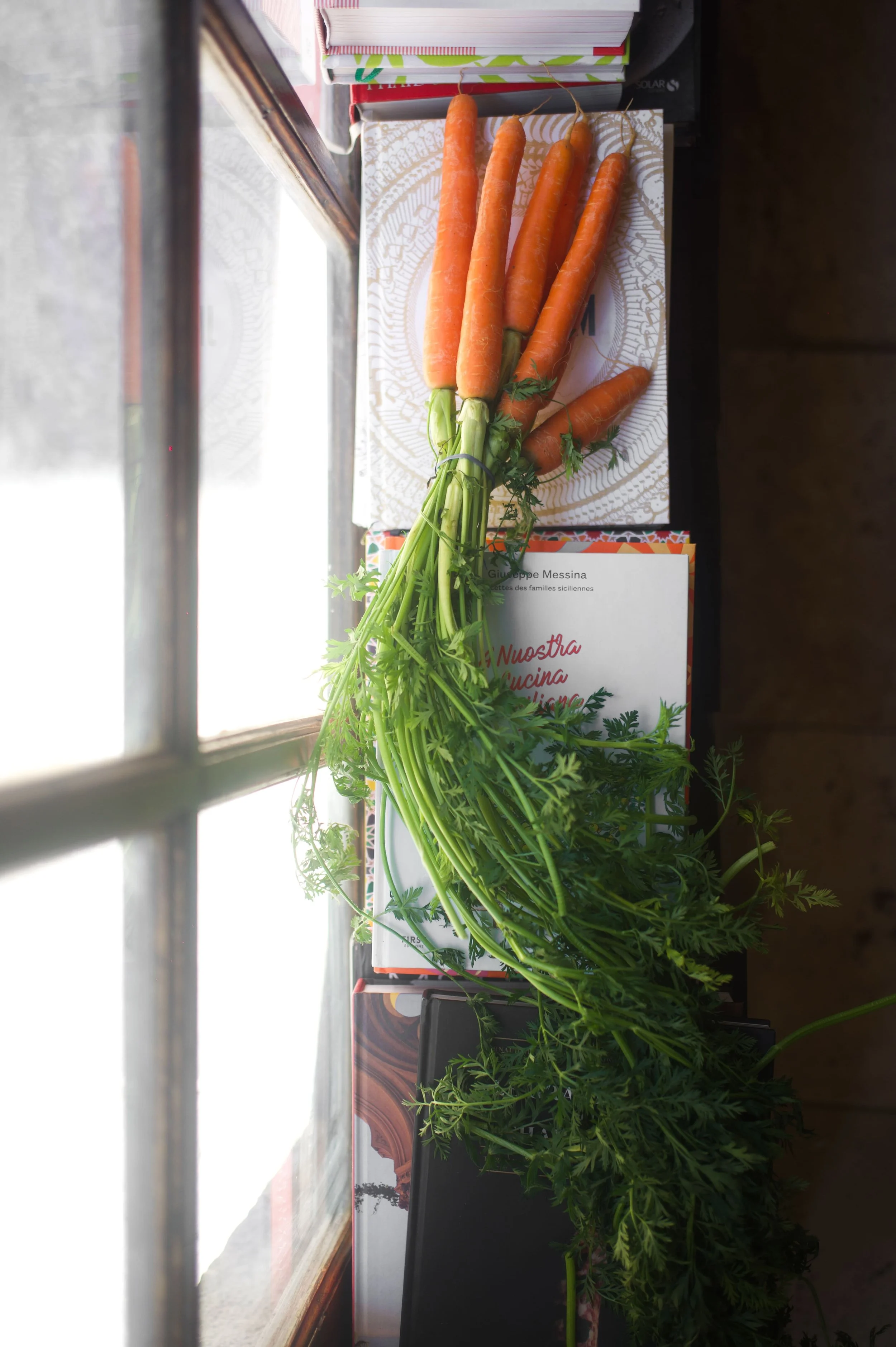Un bouquet de carottes avec feuillage vert repose sur des boîtes en carton posées sur une table près d'une fenêtre, dans une pièce lumineuse.