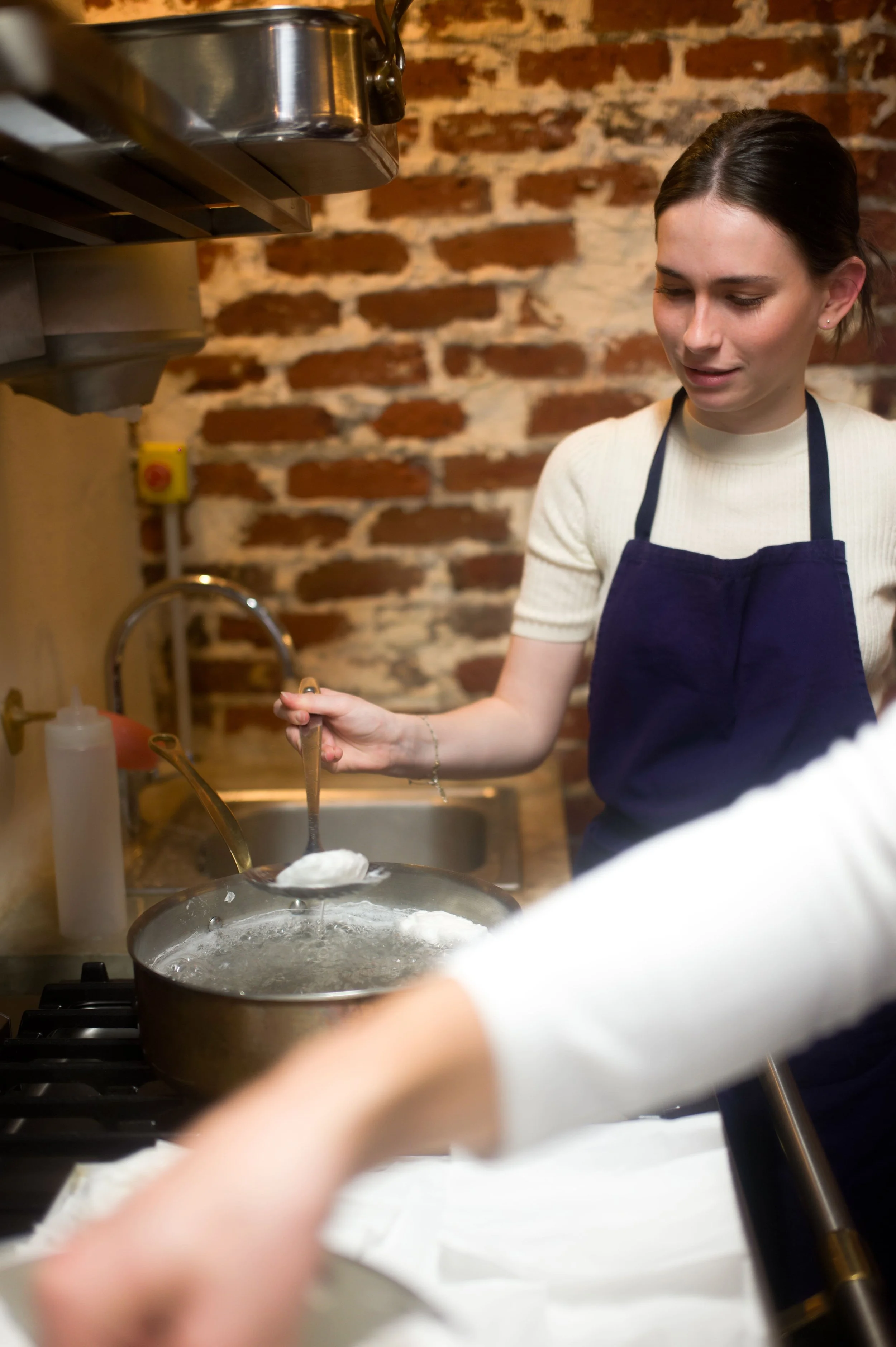 Une femme en cuisine, portant un tablier bleu et un pull blanc, utilise une écumoire pour retirer des morceaux de pâte bouillante d'une casserole à la cuisinière en cuisine.