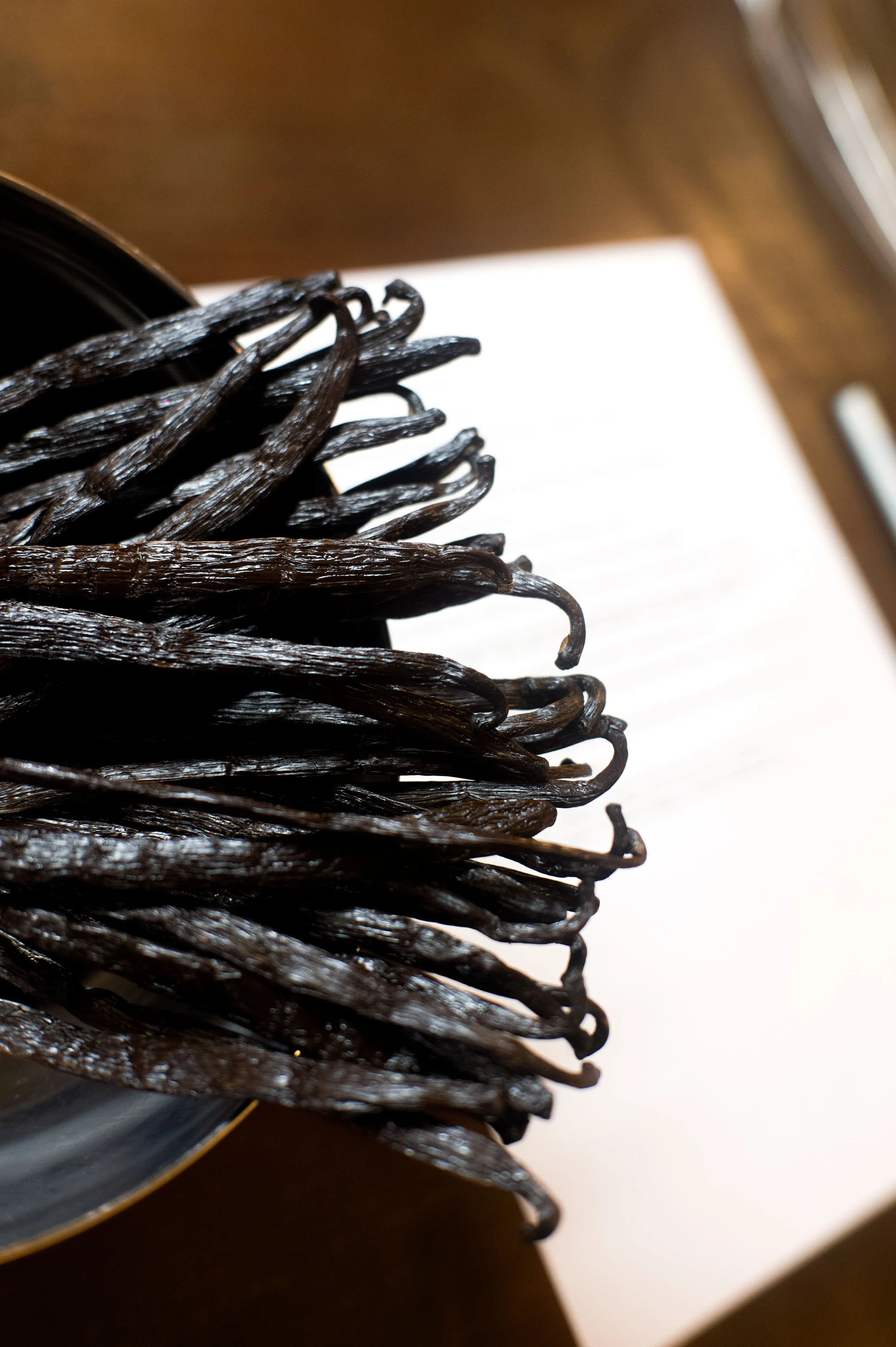 Vanille noire de tonka sur une assiette en céramique noire, posée sur une table en bois avec un carnet blanc et un stylo à côté.
