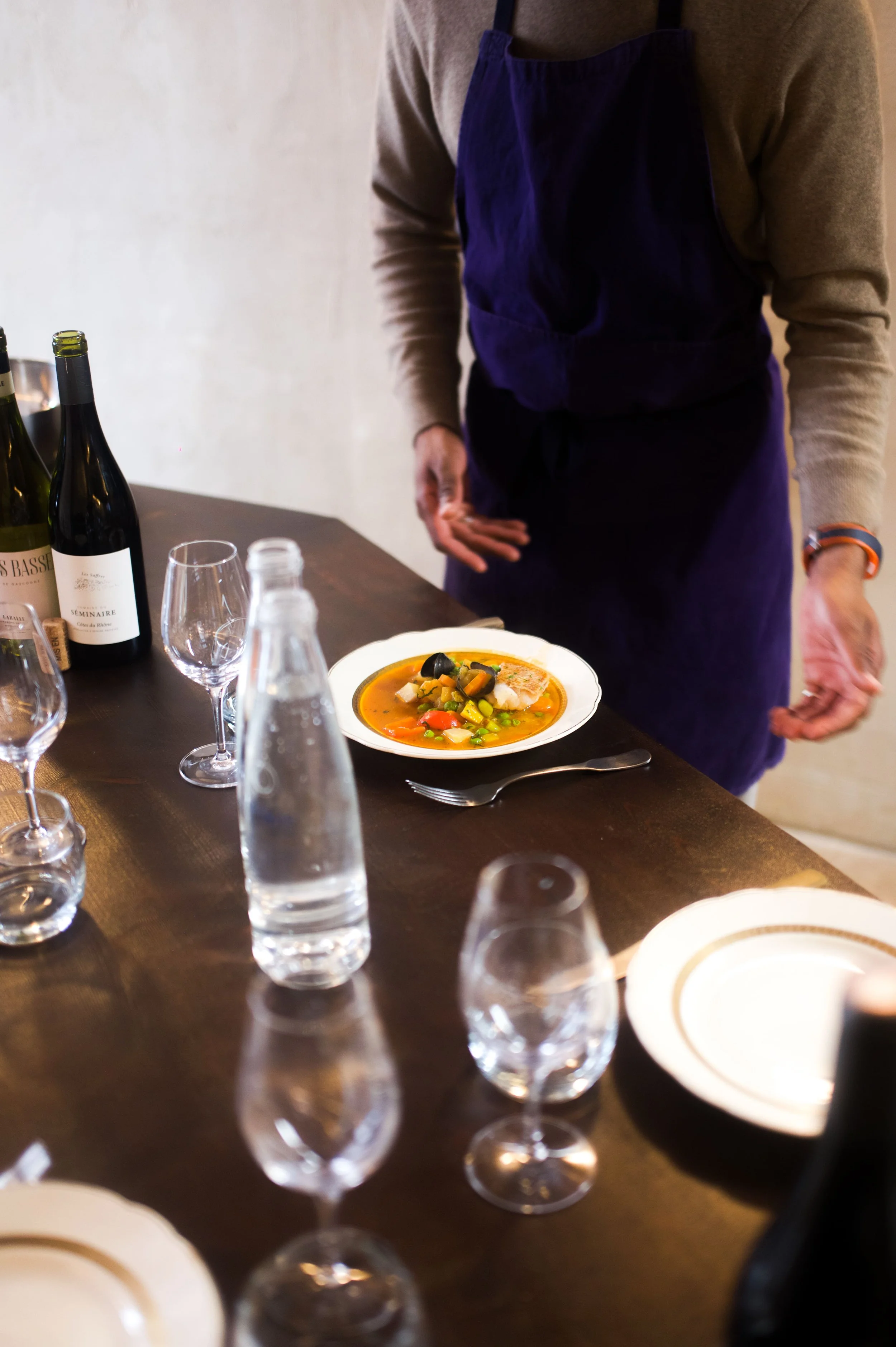Une personne en uniforme de chef présente une assiette de soupe de légumes colorée sur une table de dîner avec des bouteilles de vin, des verres à vin, une carafe d'eau et des assiettes.