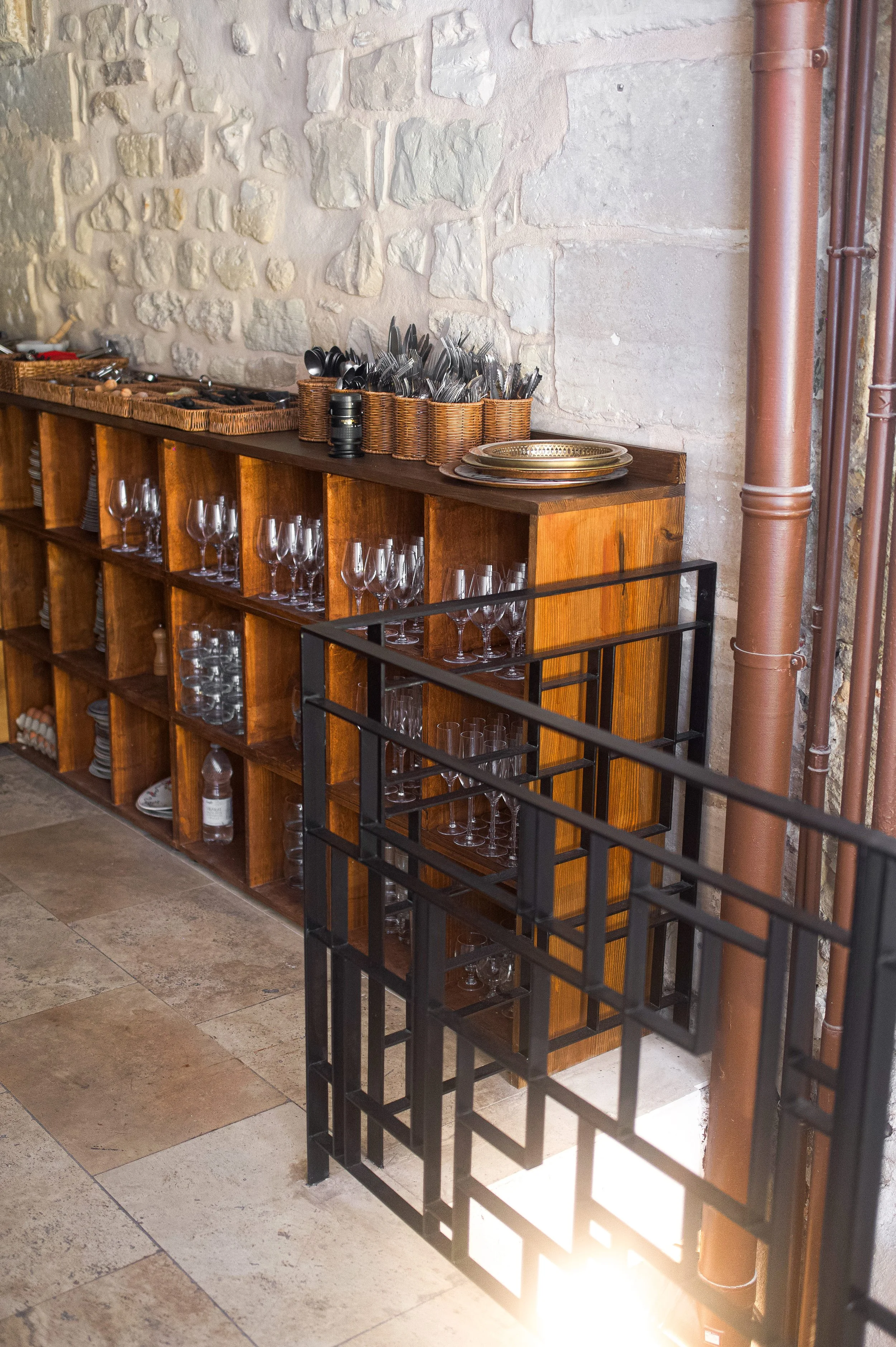 Une étagère en bois avec des verres à vin, des assiettes en métal et des ustensiles, située contre un mur en pierre dans un décor intérieur rustique.