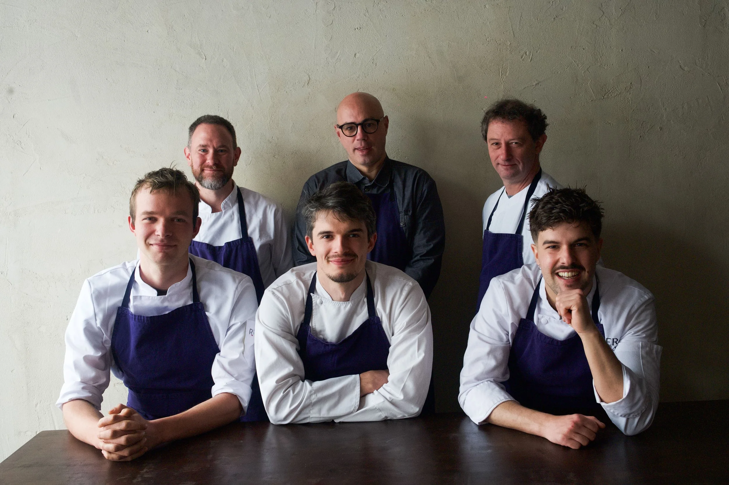 Groupe de sept chefs et cuisiniers posant debout et assis autour d'une table en cuisine, portant des uniformes blancs et des tabliers violets.