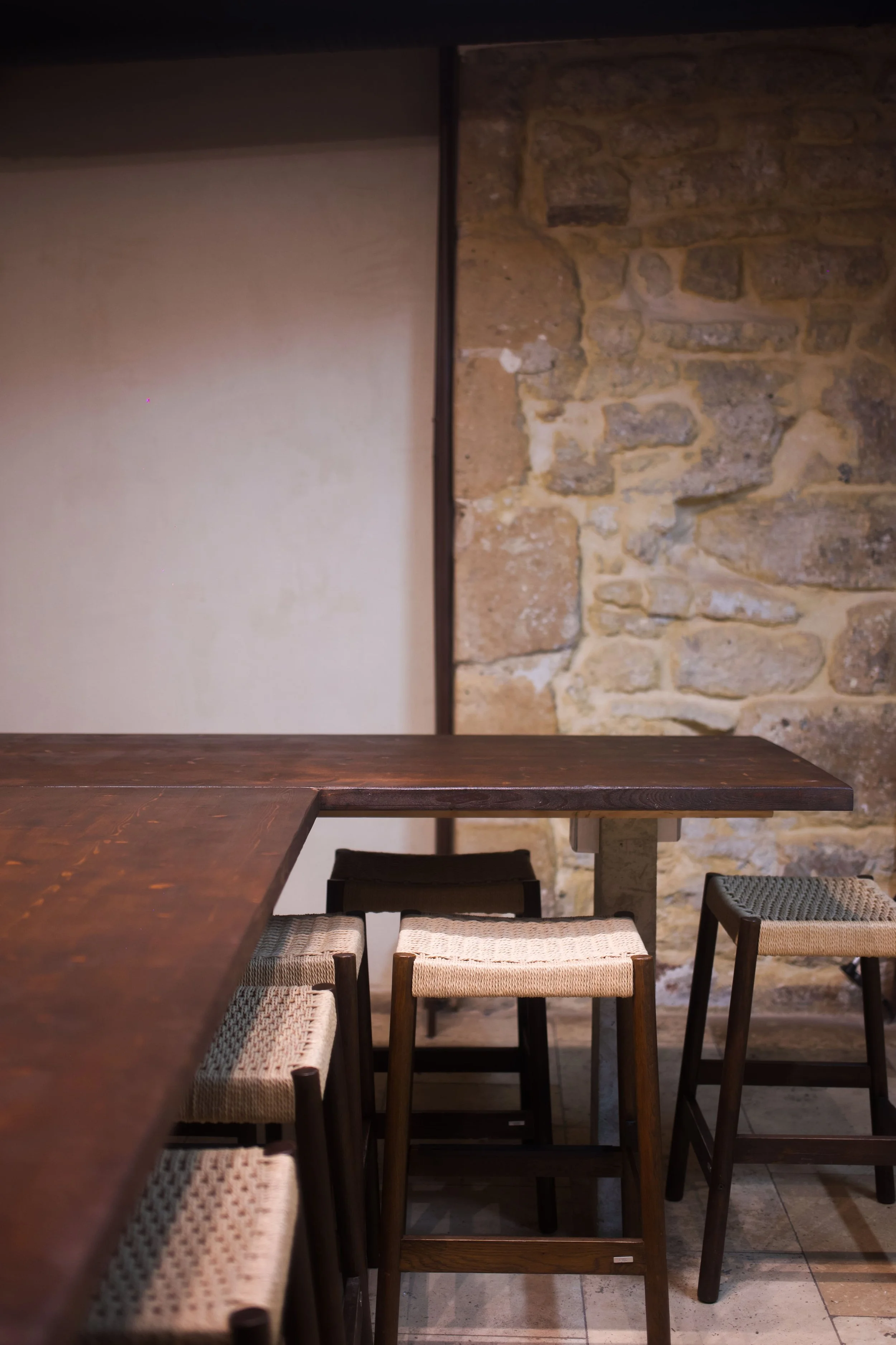 A dining area with a dark wood table, surrounded by wooden chairs with cane and fabric seats, against a stone wall and plaster partition.