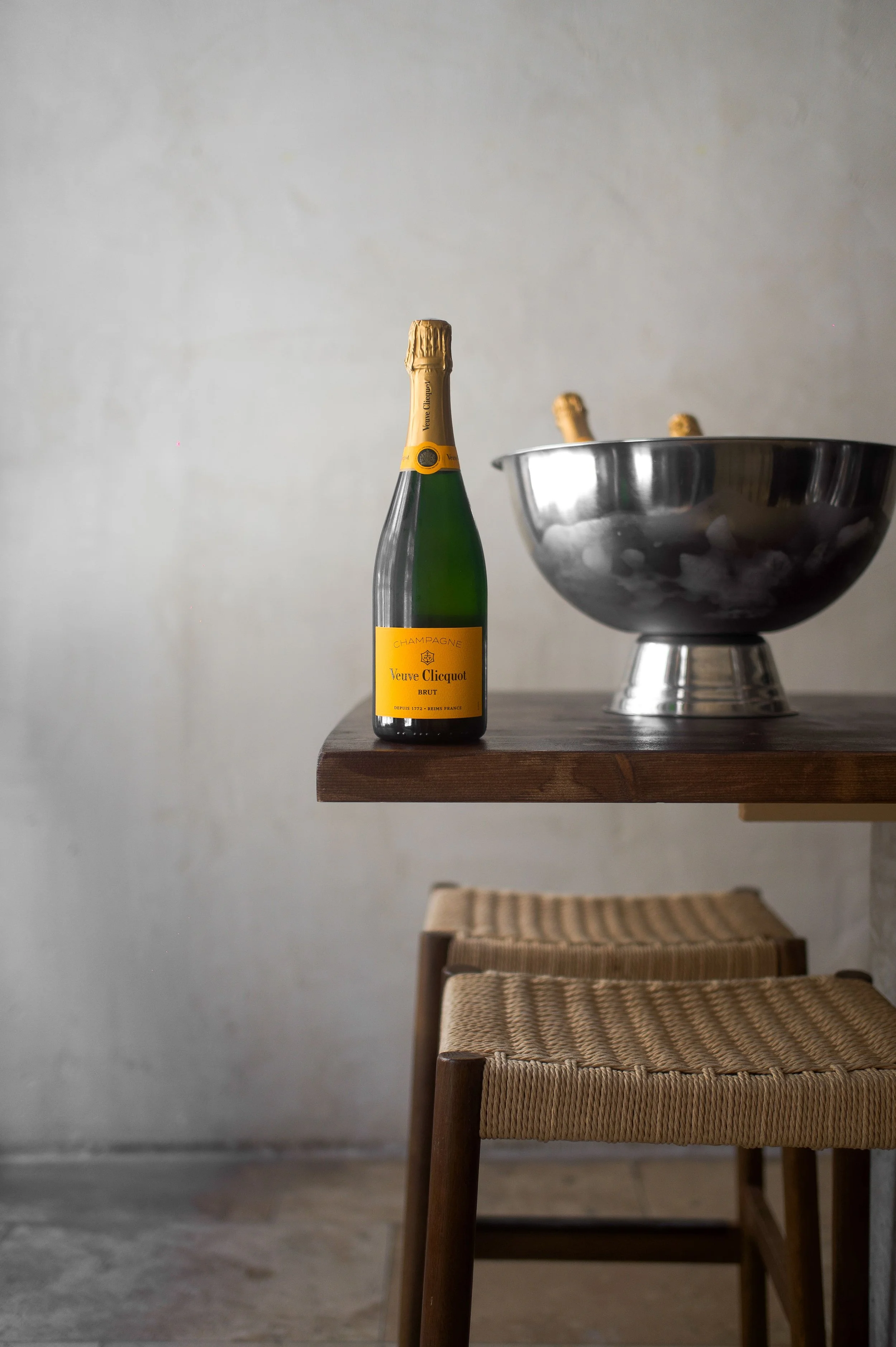 Une bouteille de champagne Veuve Clicquot sur un comptoir en bois à côté d'un bol en métal contenant des glaçons et des bouteilles de champagne. Deux chaises en bois et en corde tressée sont en dessous du comptoir.