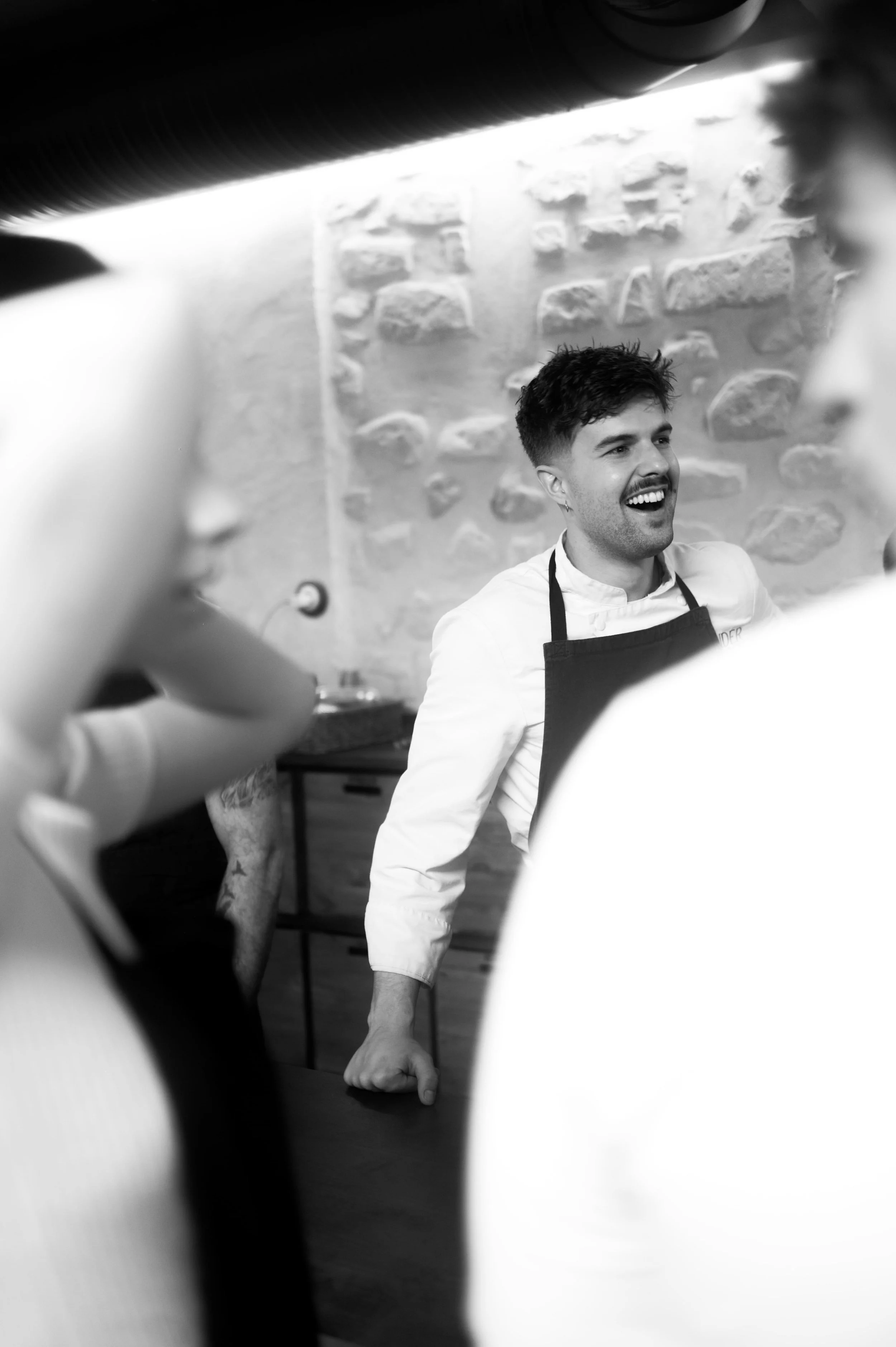 Un homme souriant, portant un tablier, entouré de personnes, dans un environnement de cuisine.