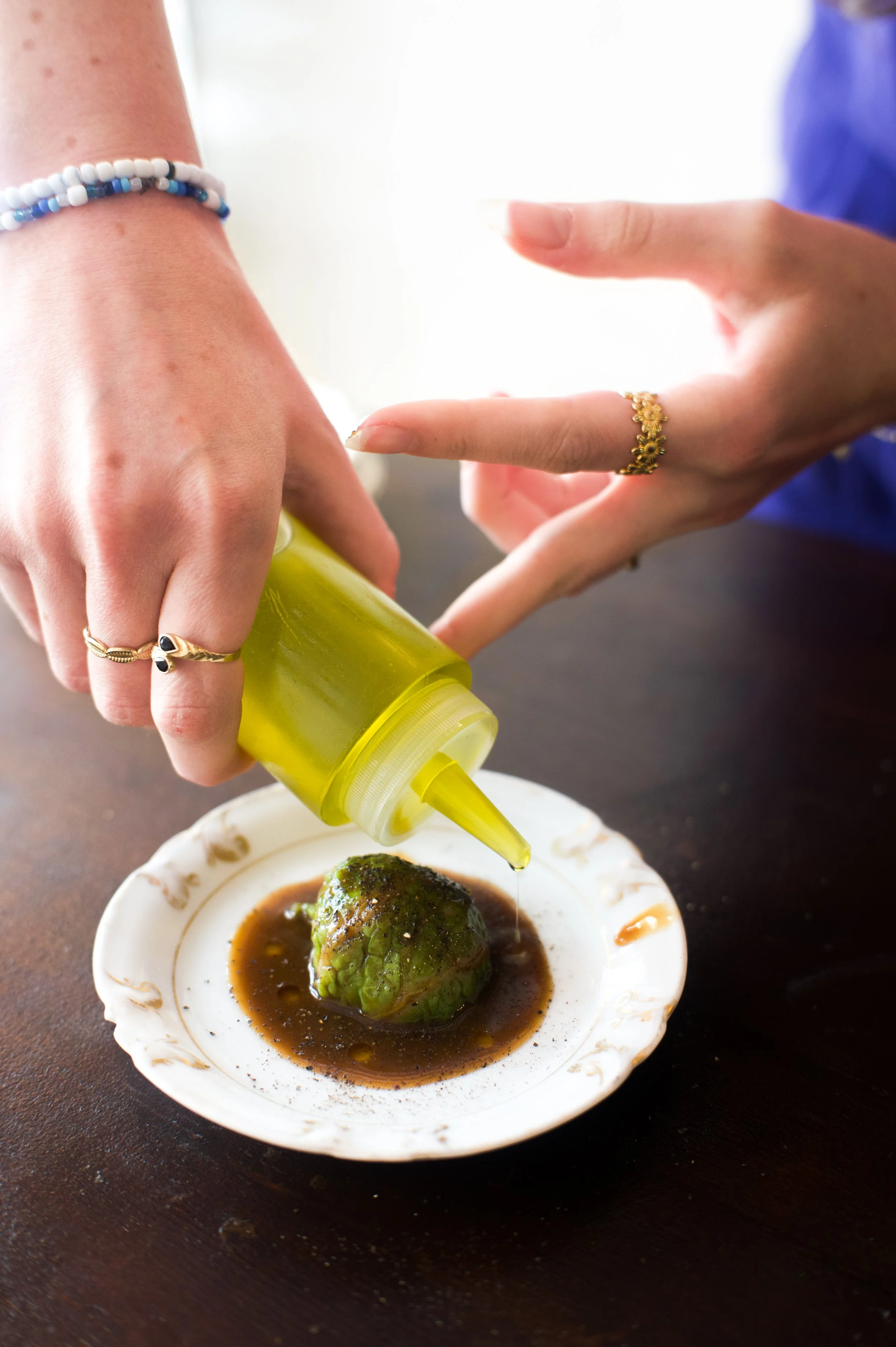 Deux mains vidangent une sauce jaune sur un morceau d'artichaut dans une assiette blanche. Les mains portent des bijoux, la scène se déroule sur une table en bois.