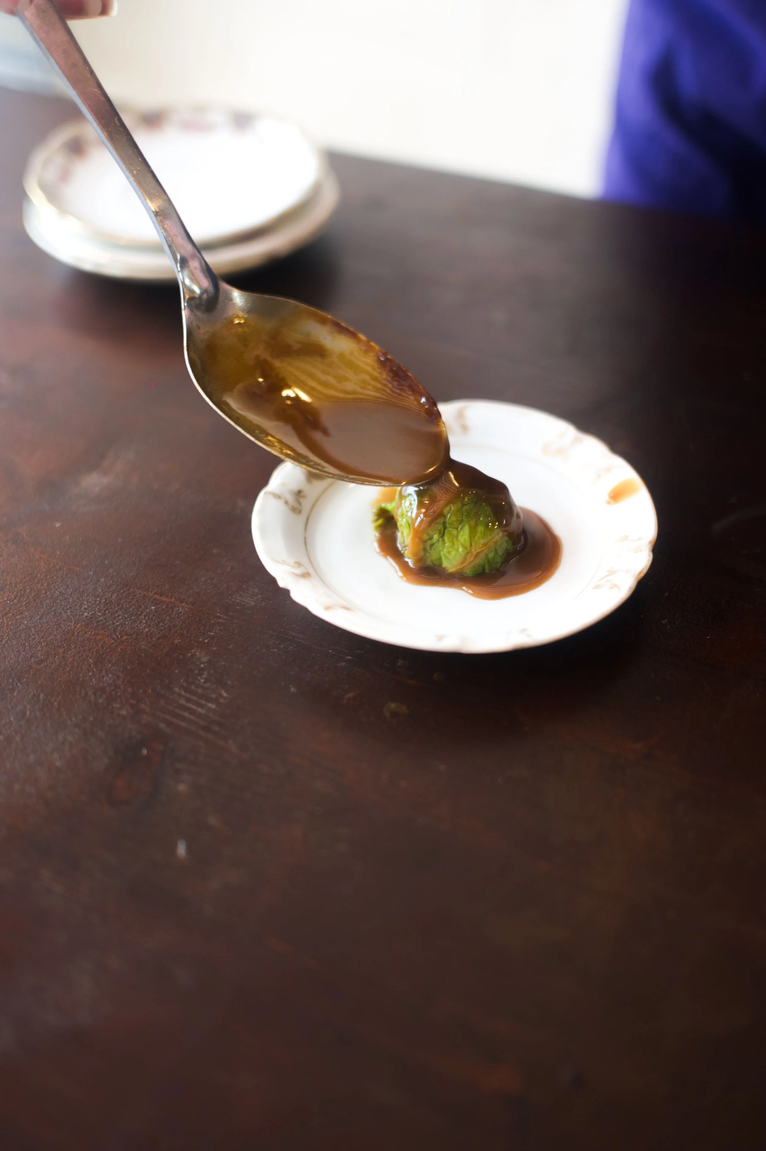 Une petite assiette blanche avec une feuille de laitue ou de salade, recouverte d'une sauce brown, sur une surface en bois. Un cuillère verse la sauce sur la feuille.