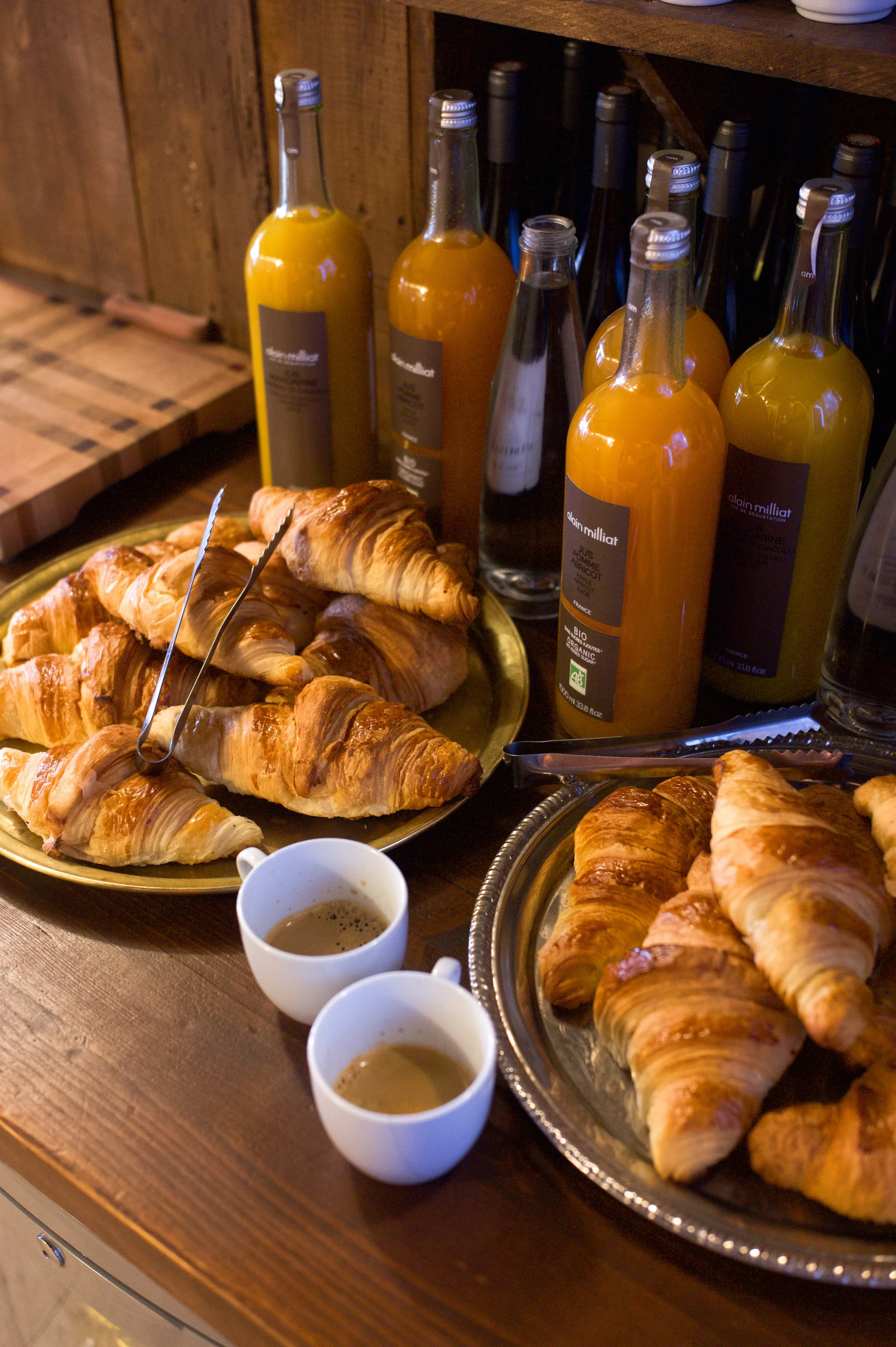 Plats de croissants frais accompagnés de bouteilles de jus d'orange et deux tasses de café sur une table en bois.