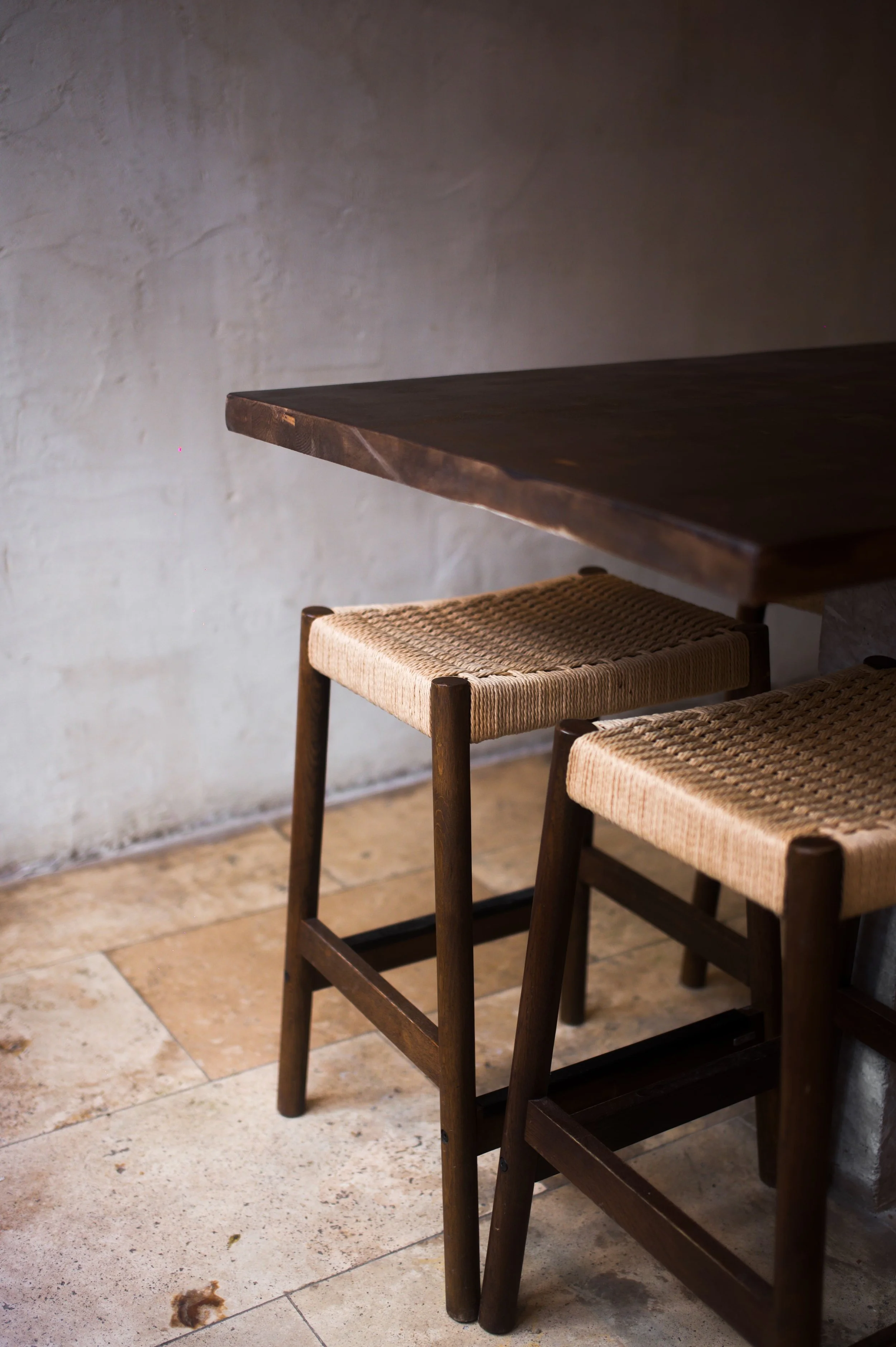 Two wooden stools with woven rope seats, placed next to a dark wooden table with a light plaster wall in the background.