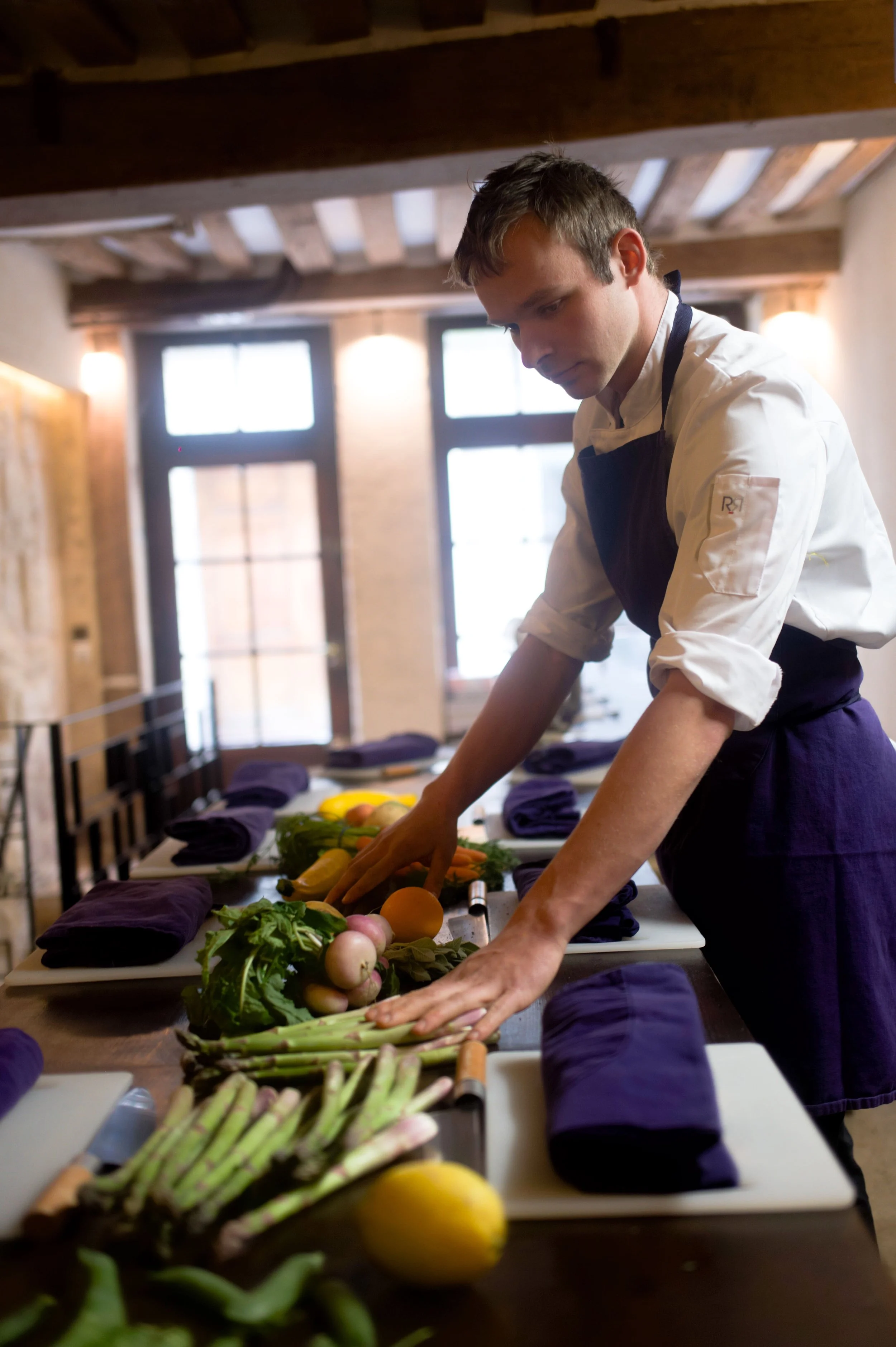 Un chef prépare des légumes frais, comprenant des asperges, des radis, des oranges et des herbes, dans une salle de cuisine en intérieur.
