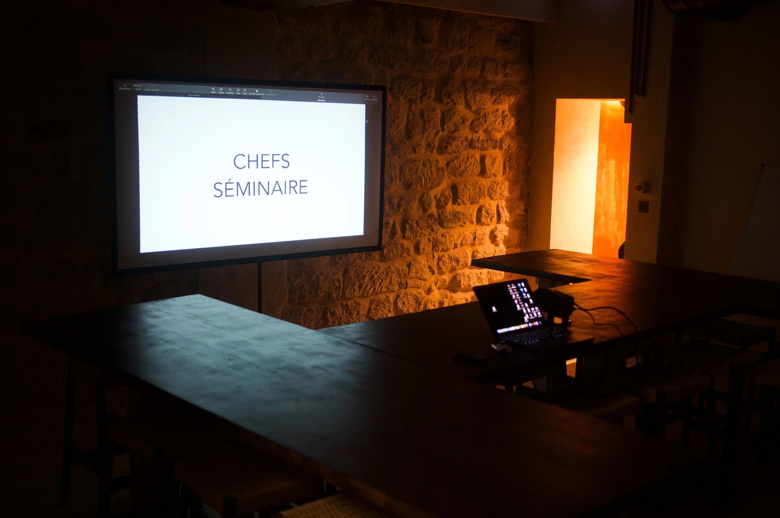 Conference room with screen displaying 'CHEFS SÉMINAIRE', wooden furniture, subdued lighting, stone wall, door ajar with light