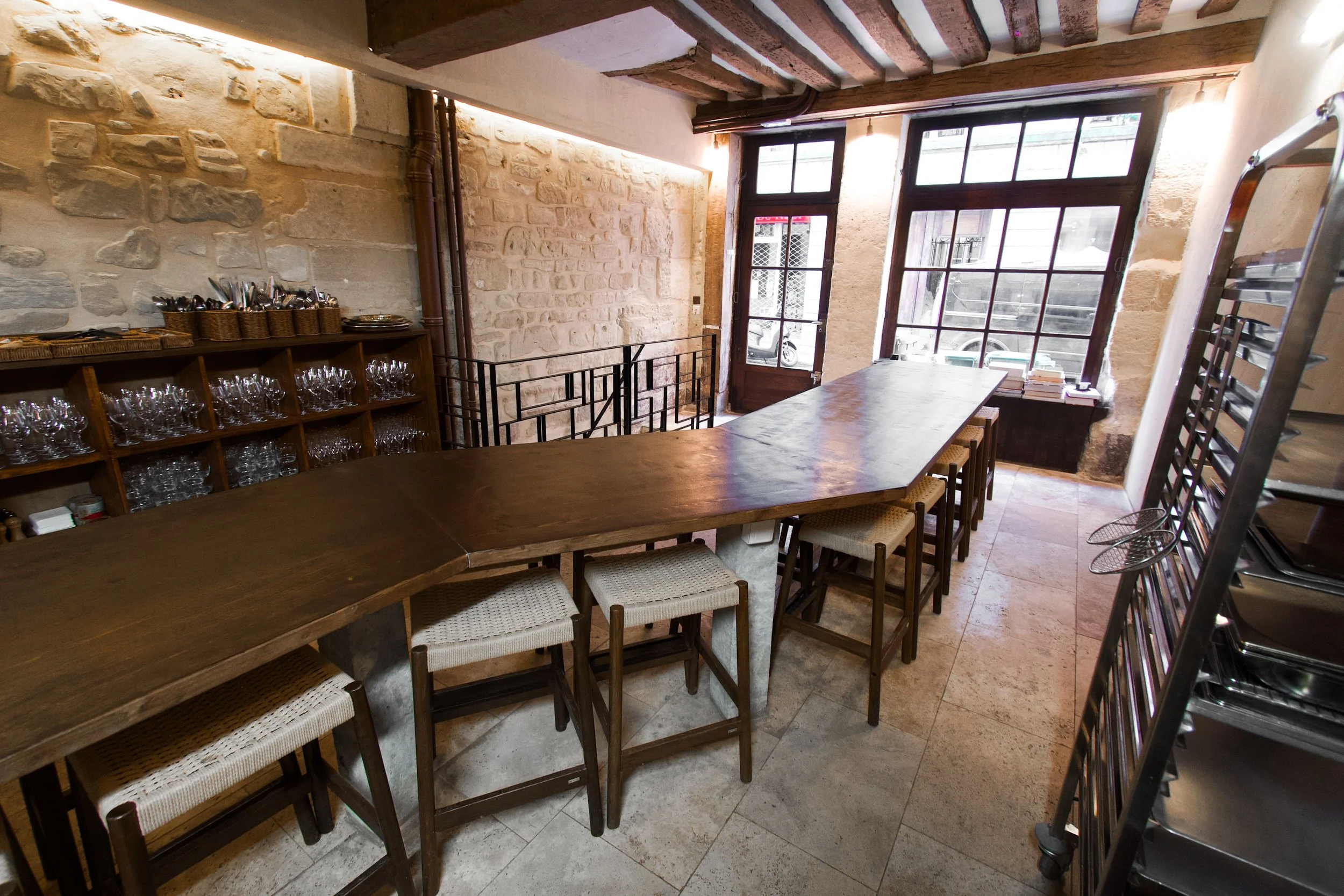 Interior of a restaurant with a large wooden island, rattan chairs, a glass shelf and large windows letting in natural light.