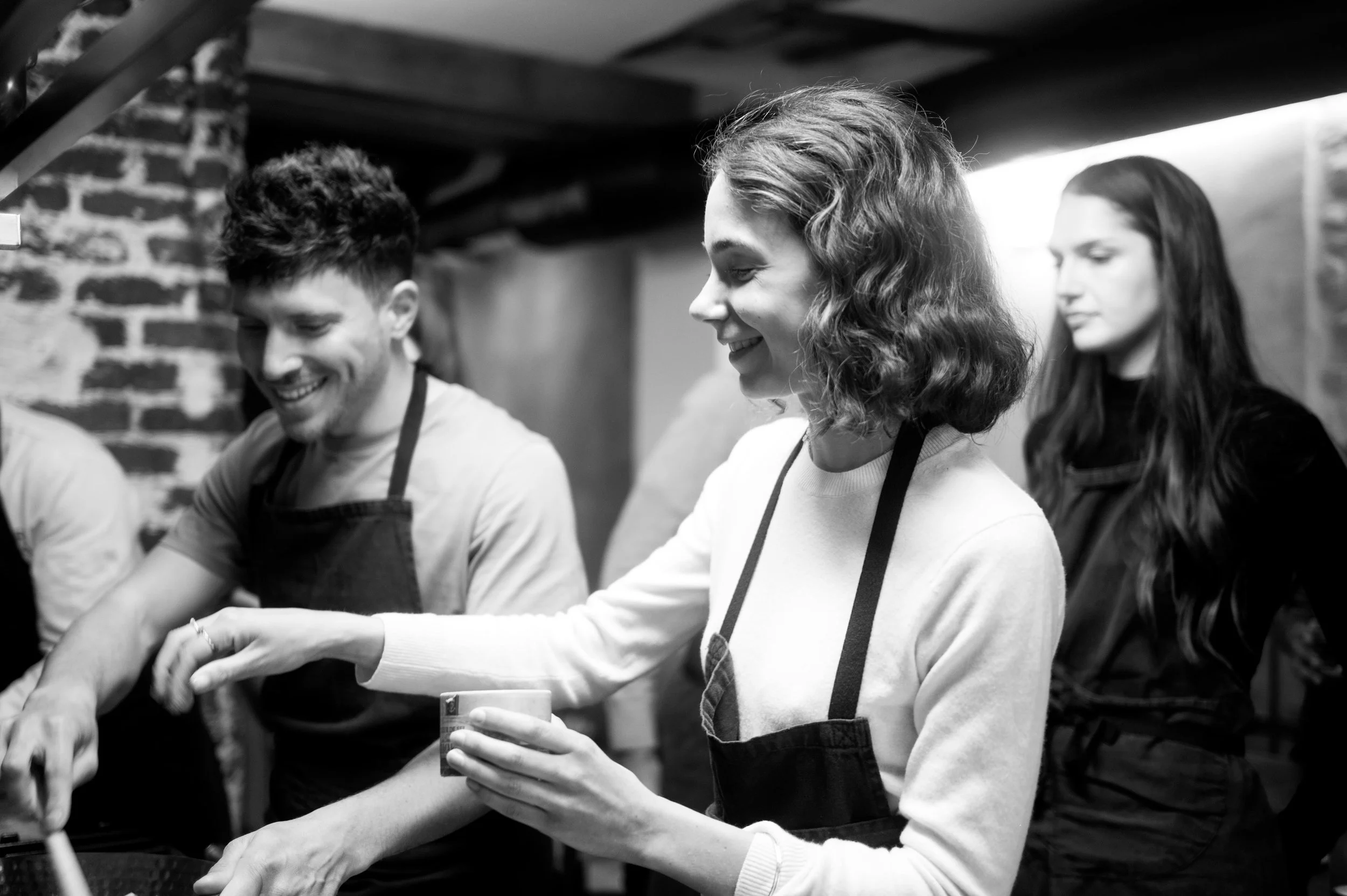 Groupe de jeunes adultes souriants préparant un repas dans une cuisine, portant des aprons.