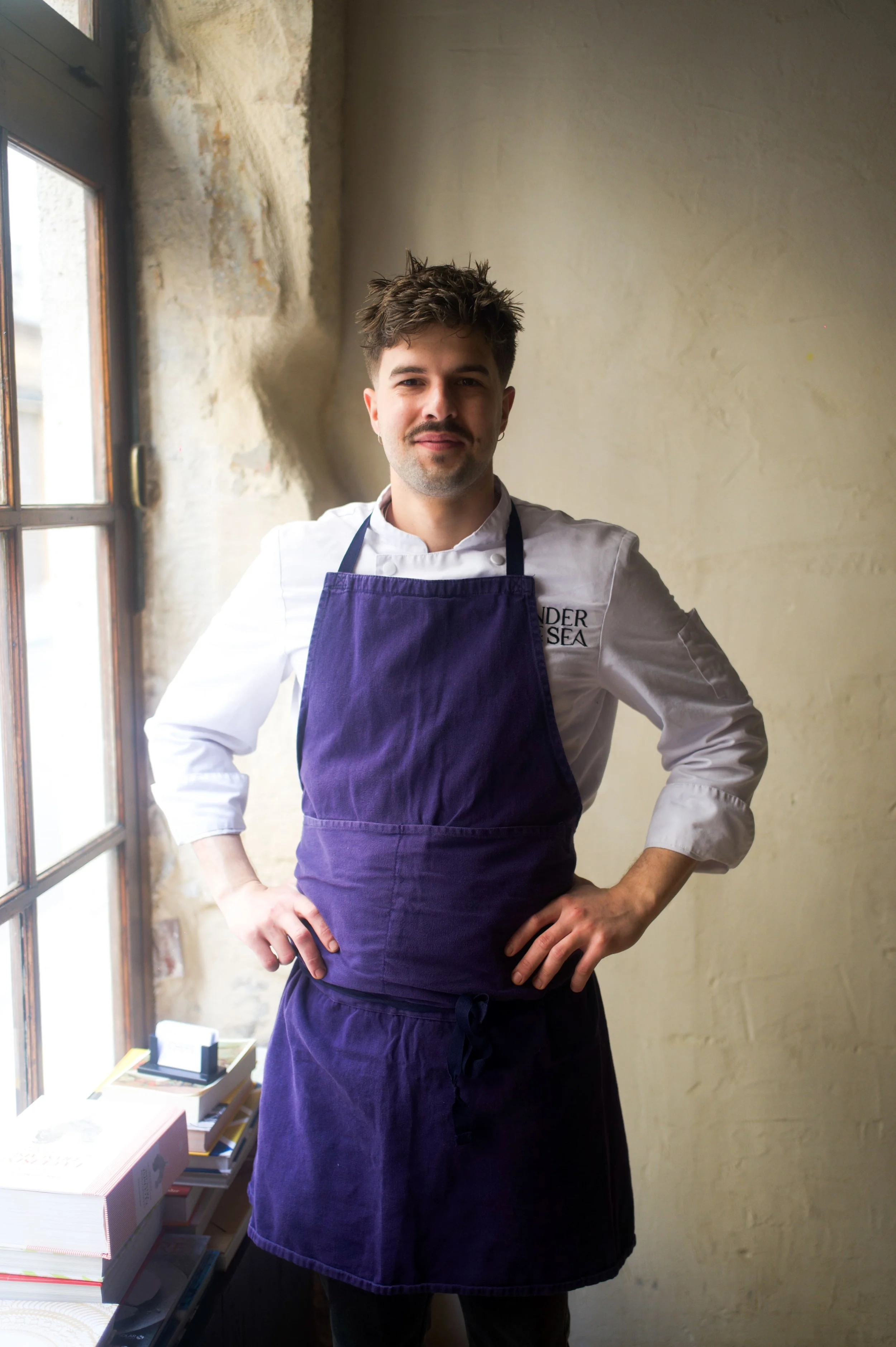 Un homme portant un uniforme de chef cuisinier, avec un tablier violet, se tient près d'une fenêtre dans une cuisine ou un restaurant.