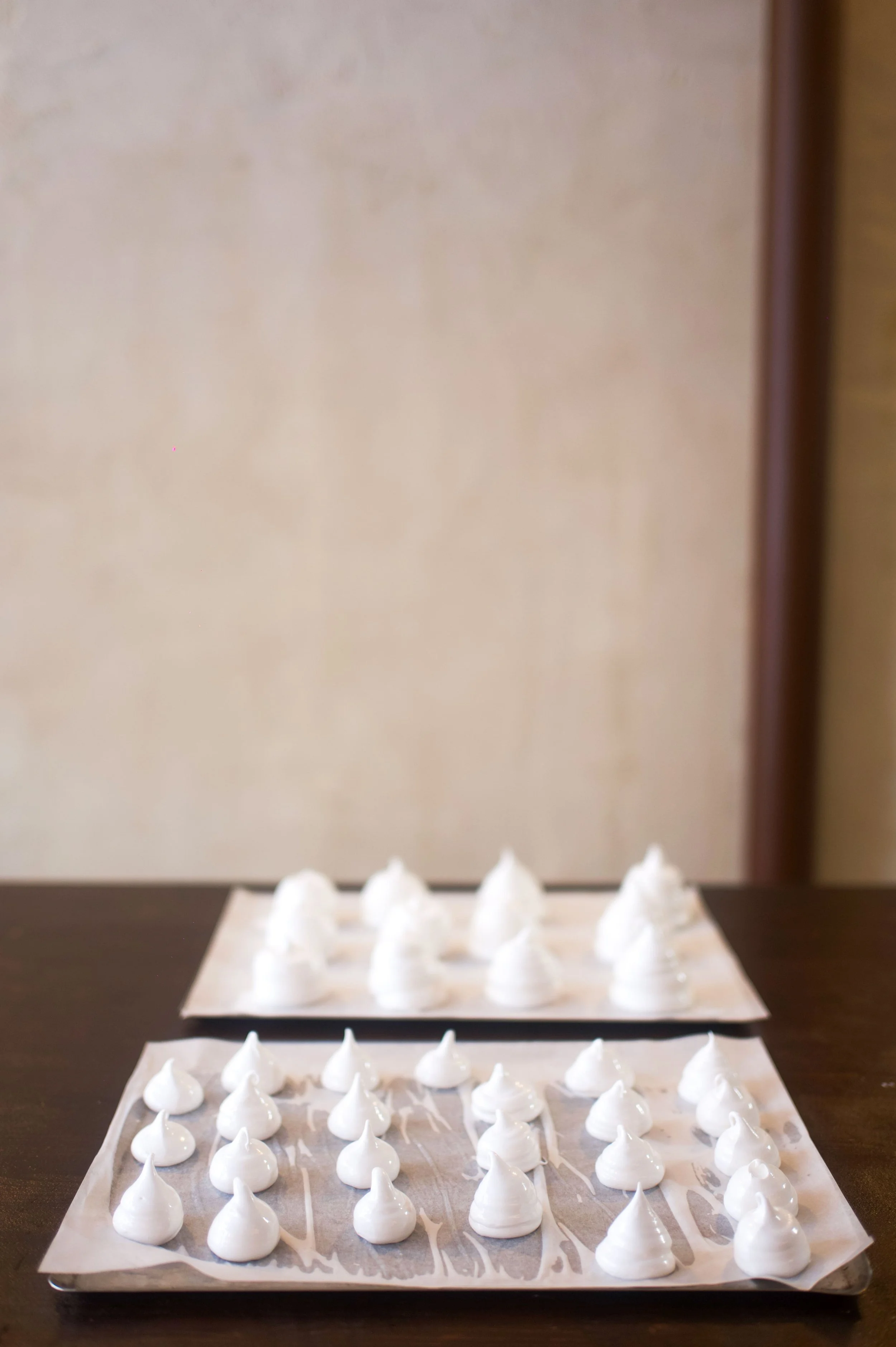 Plusieurs meringues blanches disposées sur deux plaques de cuisson recouvertes de papier sulfurisé, sur une table en bois.