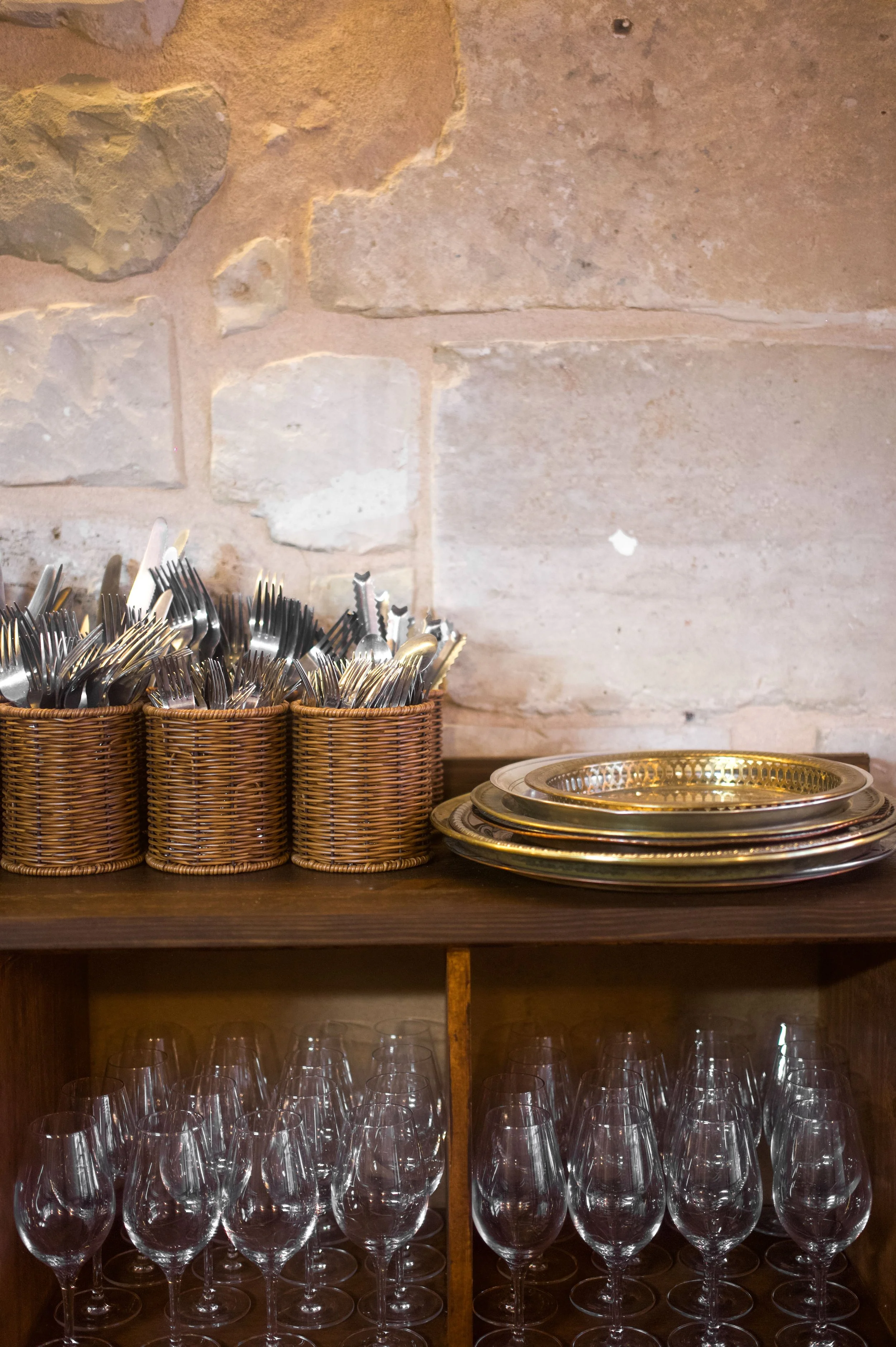 Gilded metal cutlery and plates in rattan boxes, glass wine glasses on a wooden shelf against a stone wall.