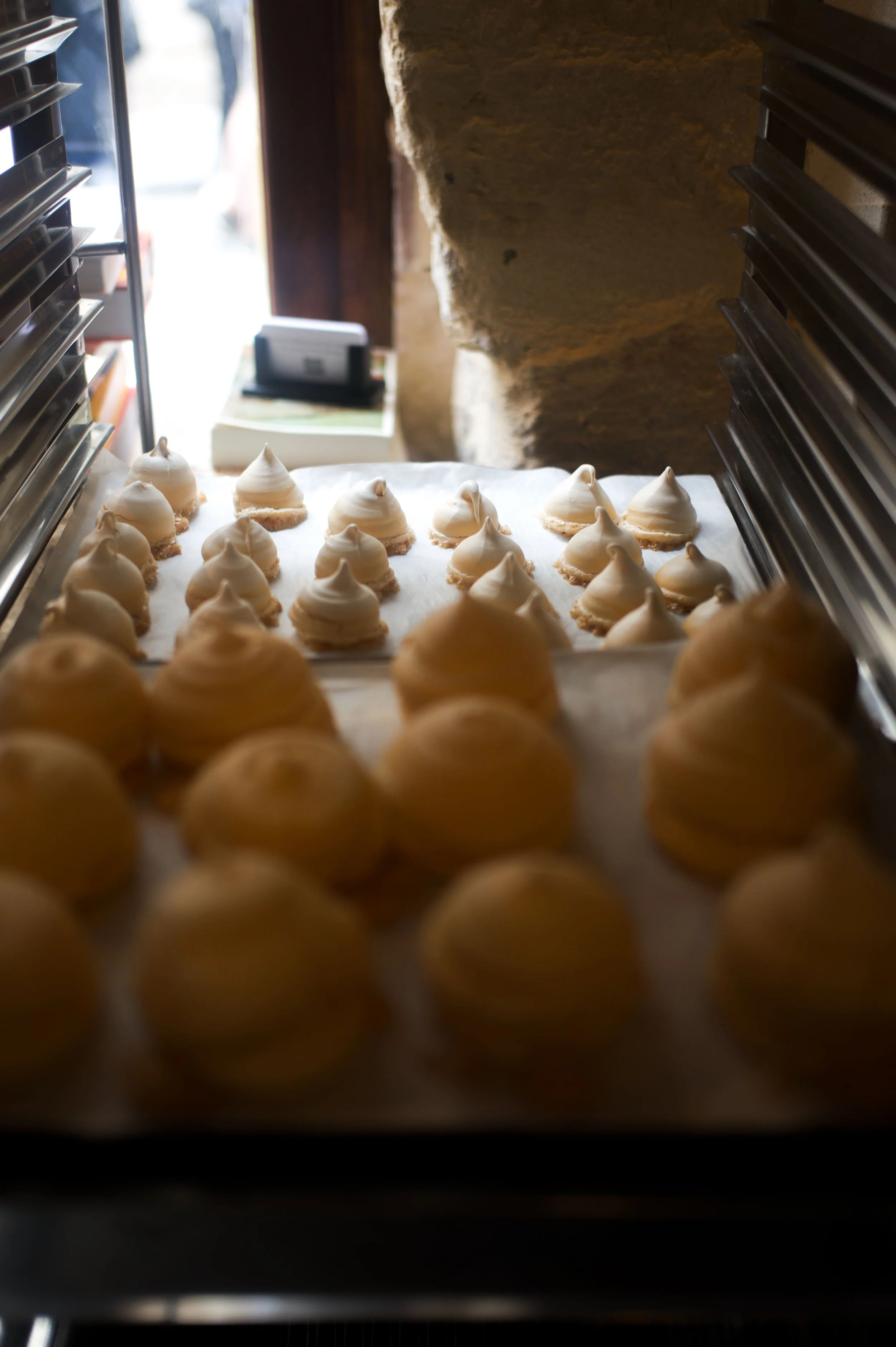 Cookies en train de cuire dans un four à pâtisserie, certains en premier plan sont flous, d'autres blancs et cuits, sur une plaque recouverte de papier sulfurisé.