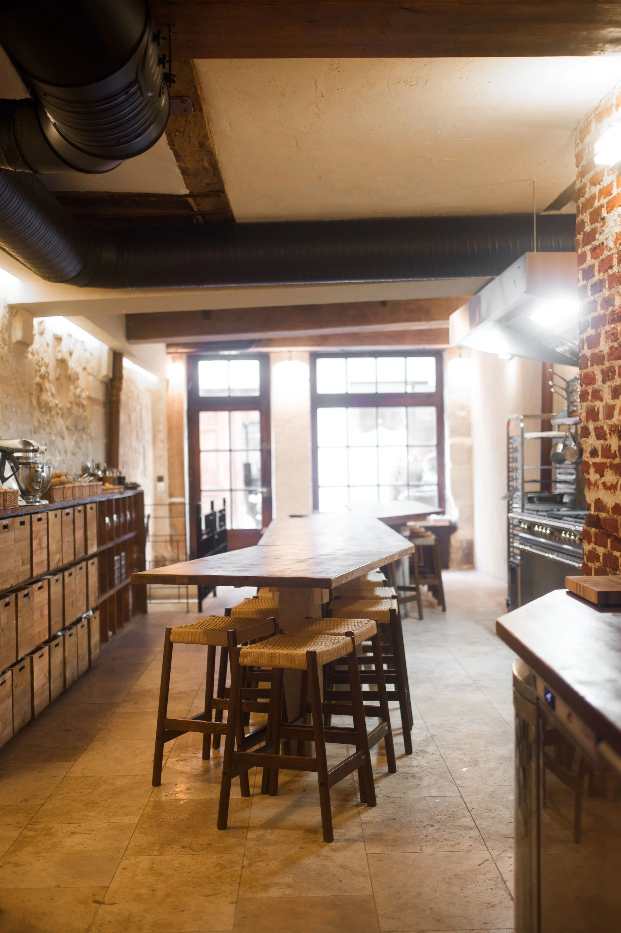 Interior of a restaurant or kitchen with a wooden counter, chairs, windows in the background, brick and stone walls, and kitchen equipment on the right.
