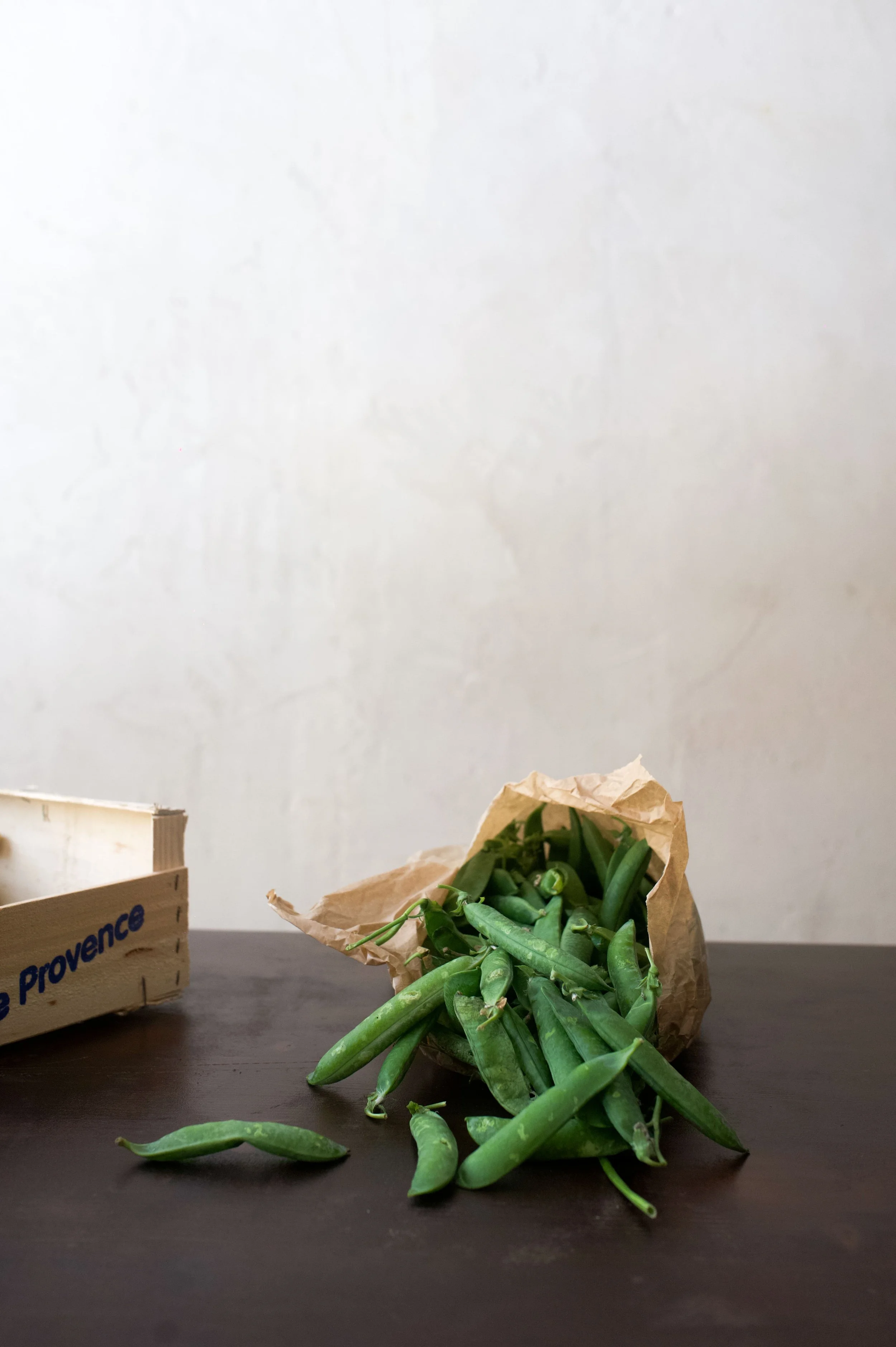 Un sac en papier débordant de gousses de pois verts sur une table en bois, avec une boîte en bois partiellement visible à gauche.