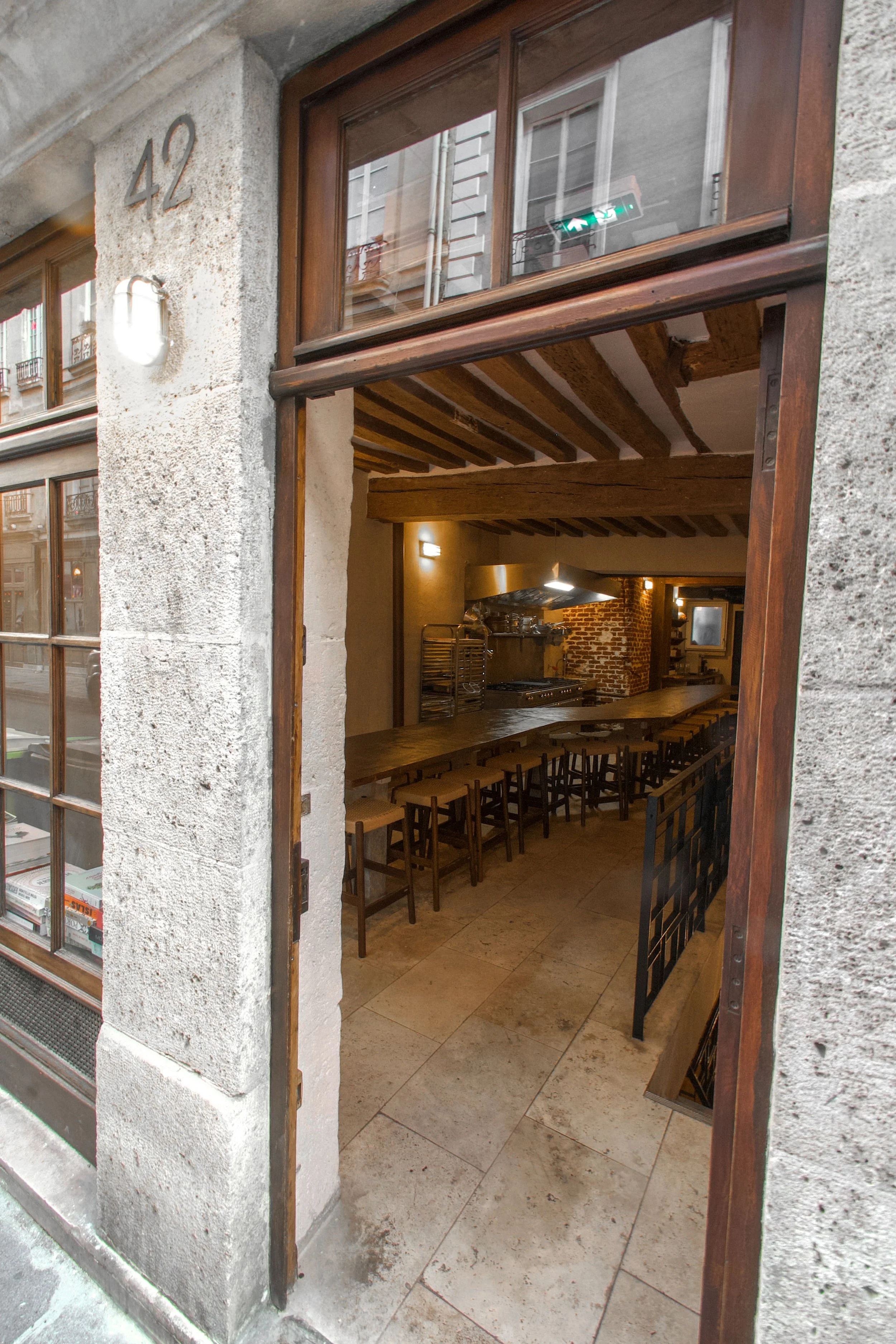 View of a restaurant entrance with a large open window, showing a warm interior with a long wooden table, chairs, wood-beamed ceilings and an exposed brick wall.
