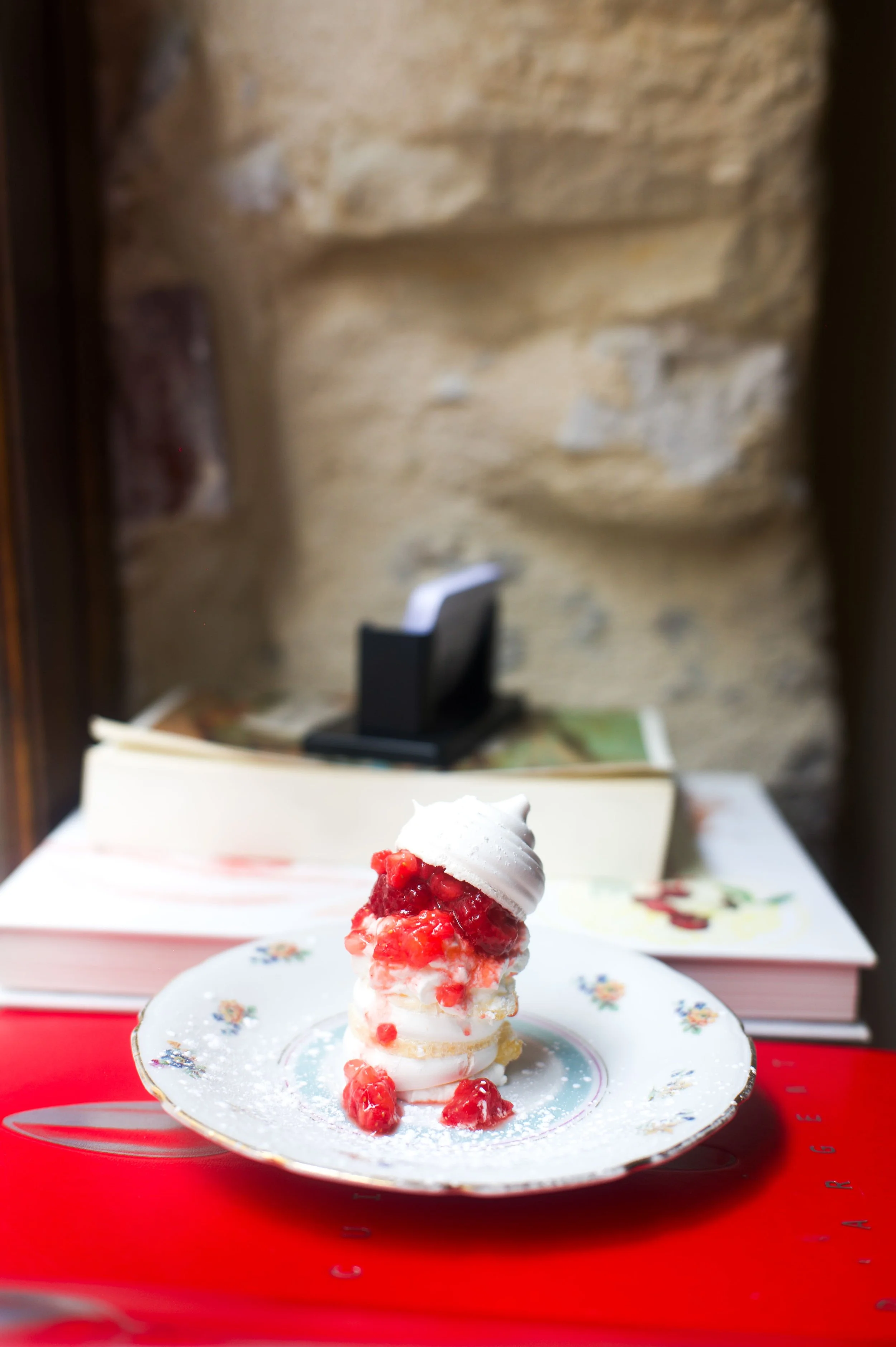 Une pavlova avec des fruits rouges, crémée, sur une assiette décorative blanche, posée sur une surface rouge, avec des livres en arrière-plan et un mur en pierre.