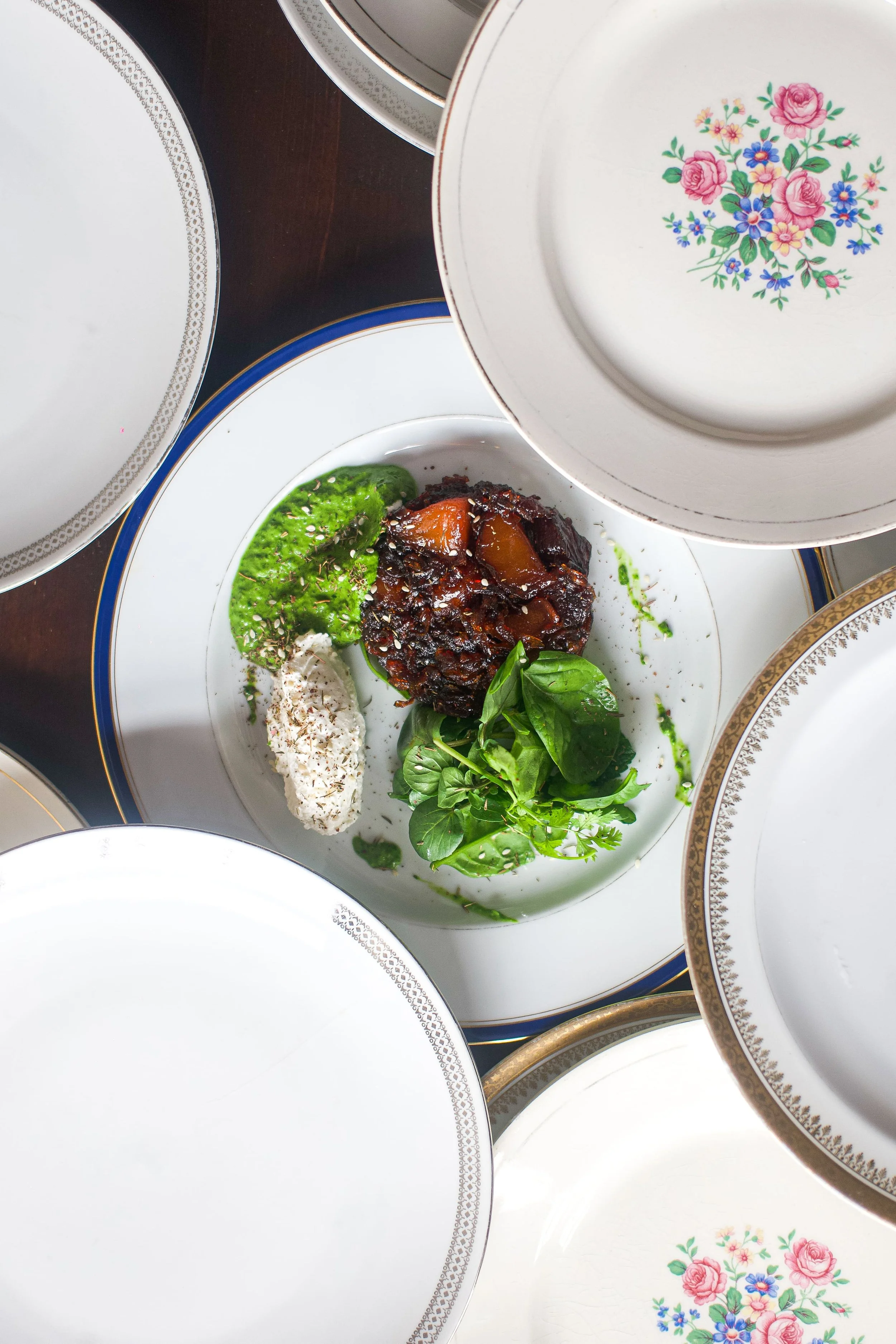 Assiette avec un plat de viande en sauce, accompagnée de légumes verts et de crudités, entourée de plusieurs assiettes décoratives en vaisselle ancienne.