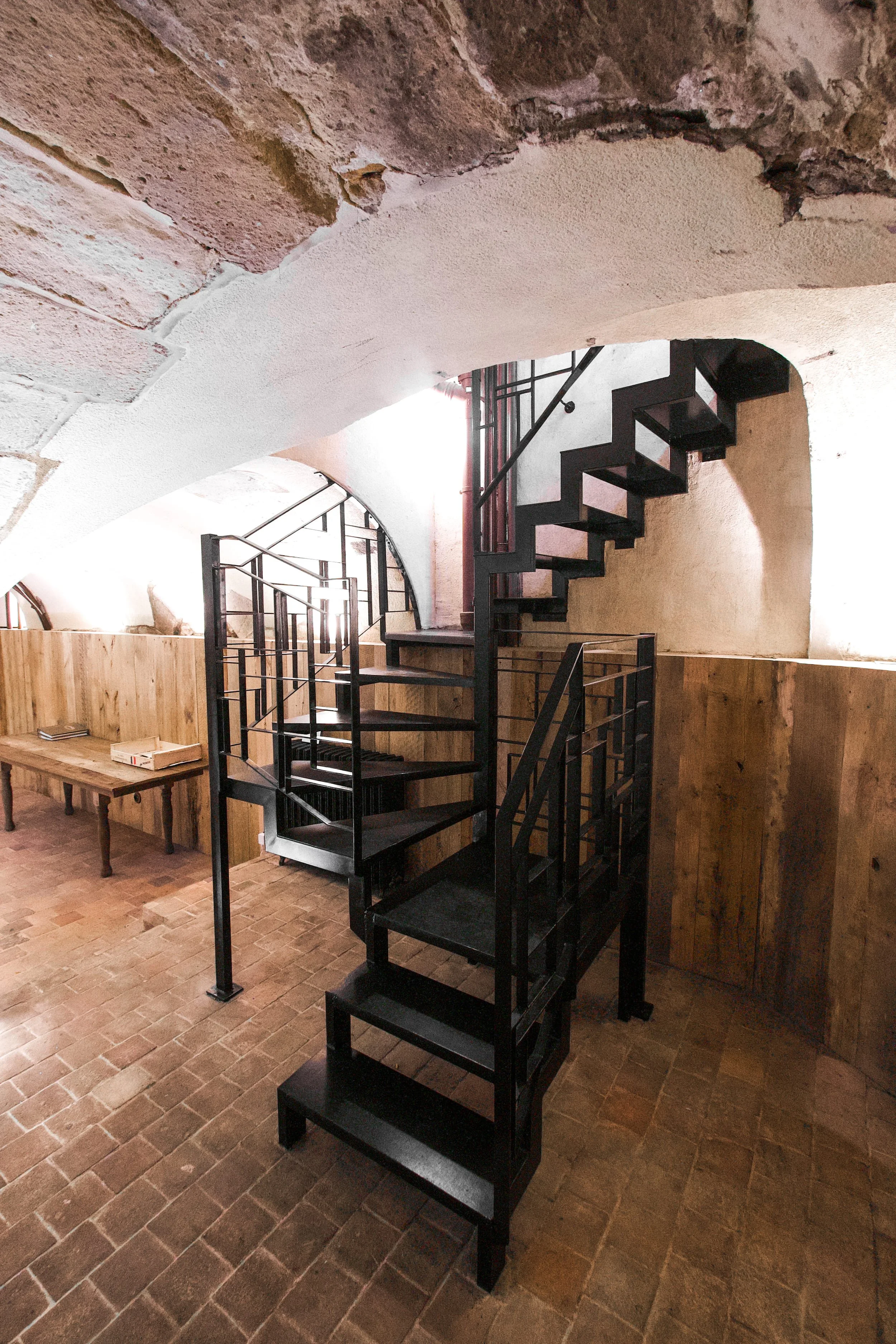 Black metal lightning bolt staircase in a room with stone walls, vaulted stone ceiling, terracotta tile floor and wooden furniture.