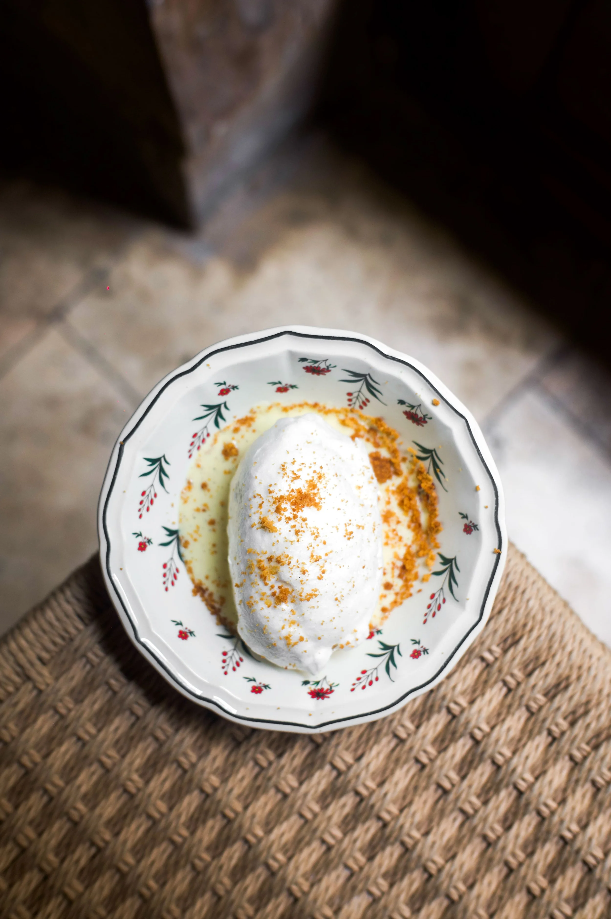Une assiette décorative contenant un dessert blanc, probablement une mousse ou une panna cotta, garni d'une poudre orange (probablement des biscuits écrasés) sur une surface en bois tressé.