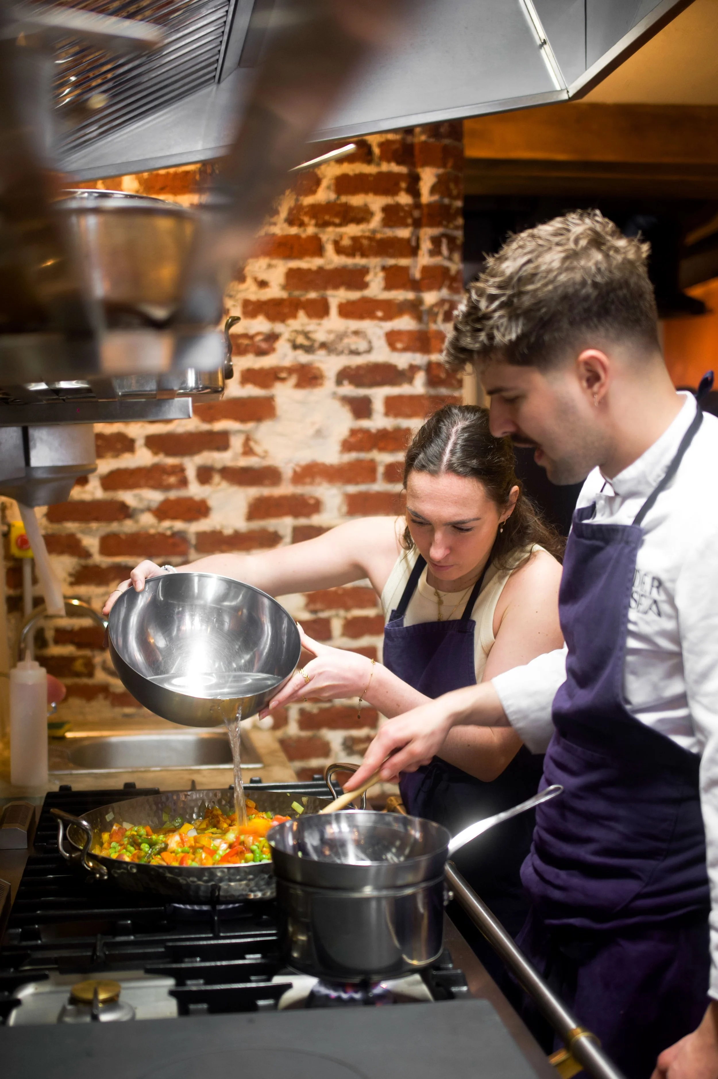 Deux chefs cuisiniers en train de préparer un plat dans une cuisine professionnelle, avec un mur en briques en arrière-plan.