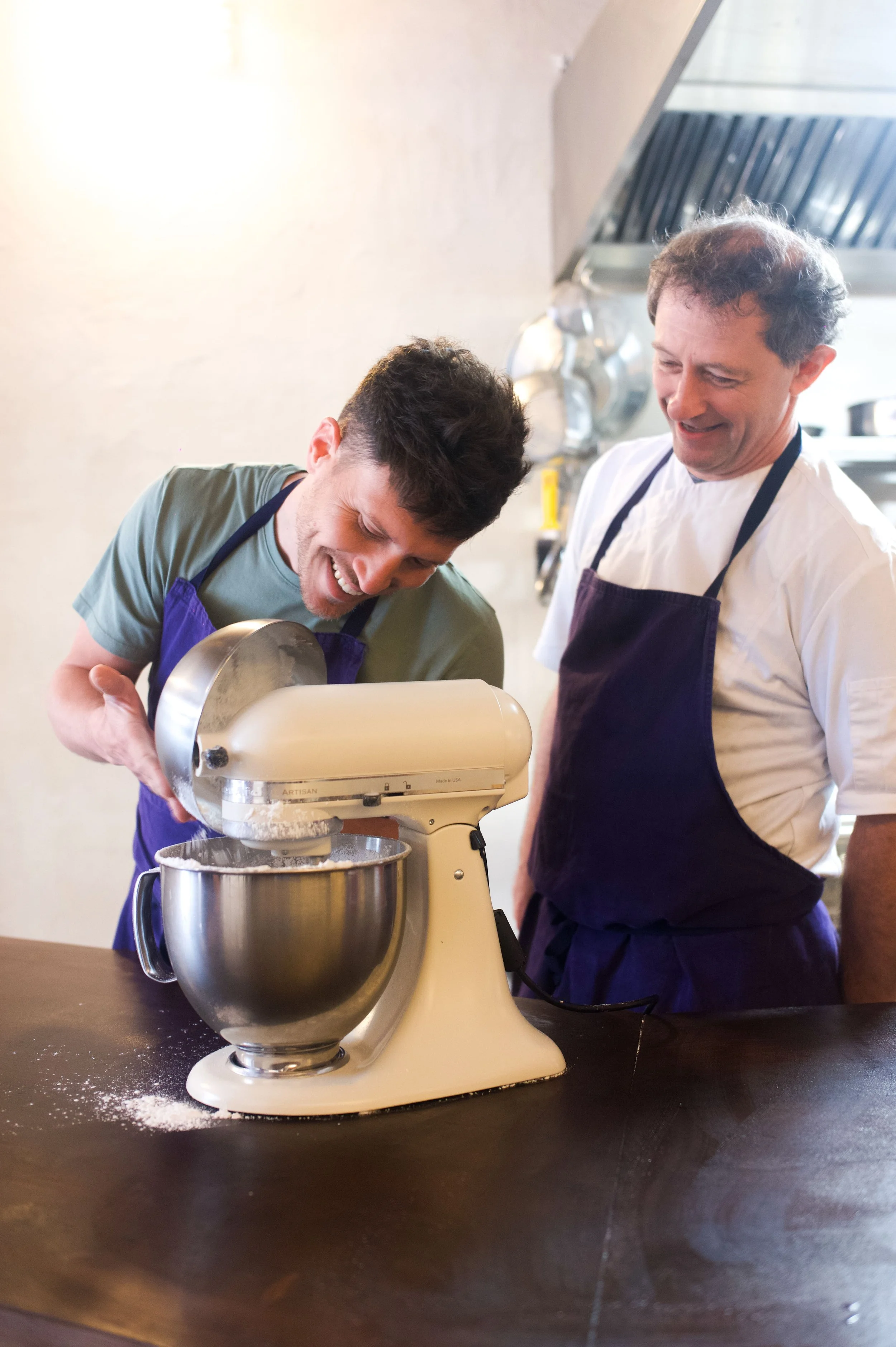 Deux hommes en tabliers de cuisine, un utilisant un robot pâtissier pour mélanger de la farine, tous deux souriants, dans une cuisine