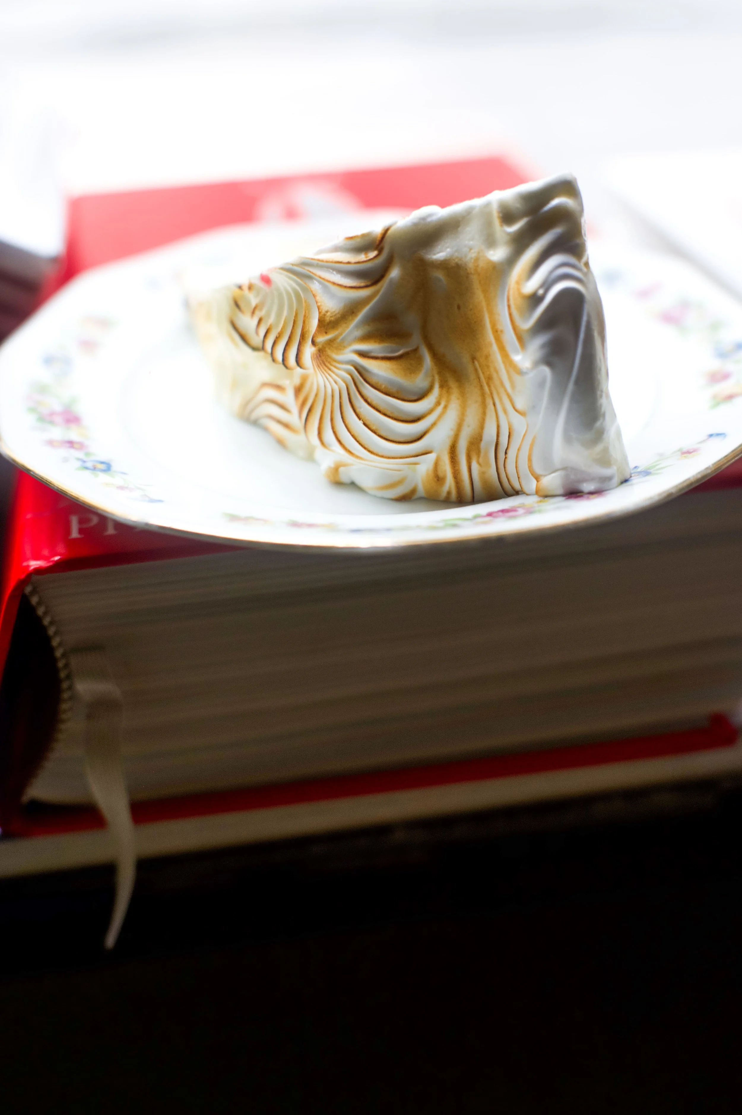 Une part de tarte à la crème avec un décor de caramel, posée sur une assiette en porcelaine posée sur un livre à couverture rouge.